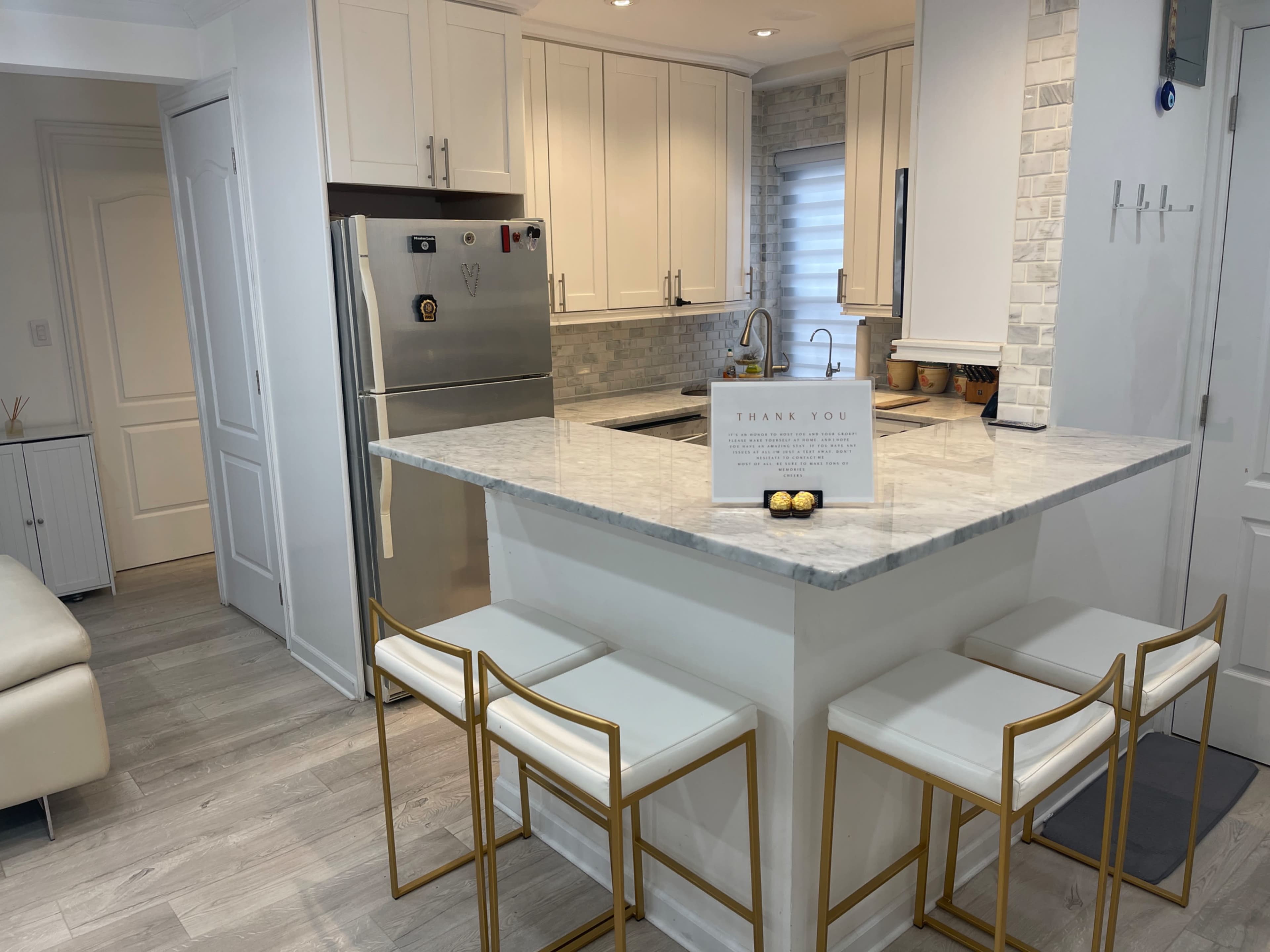 A modern kitchen features a marble countertop island with three gold-accented stools, a stainless steel refrigerator, and light-colored cabinetry.
