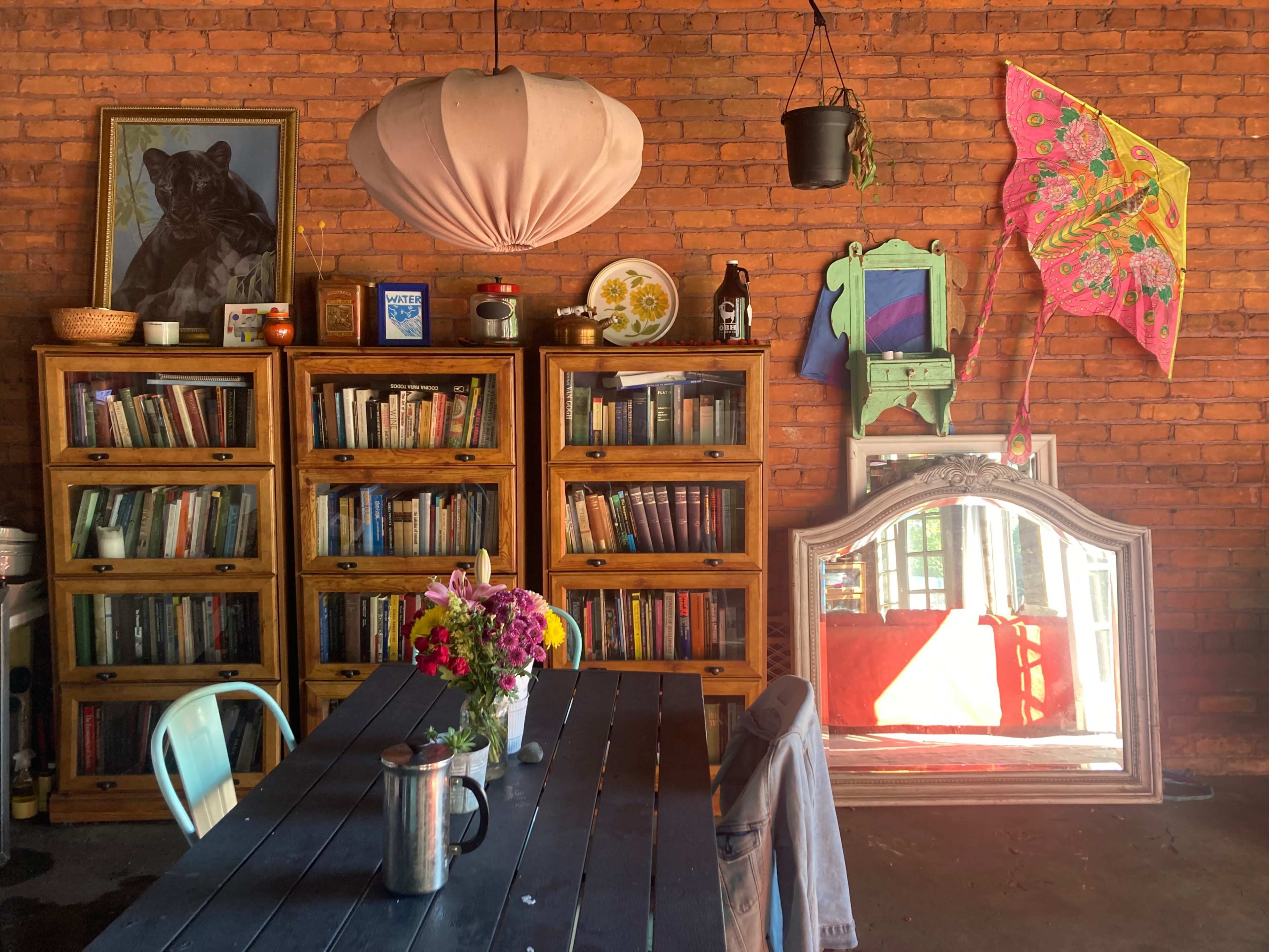 A cozy interior features wooden bookshelves filled with books, a table adorned with flowers, and colorful decor including a large mirror and a paper kite against a brick wall.
