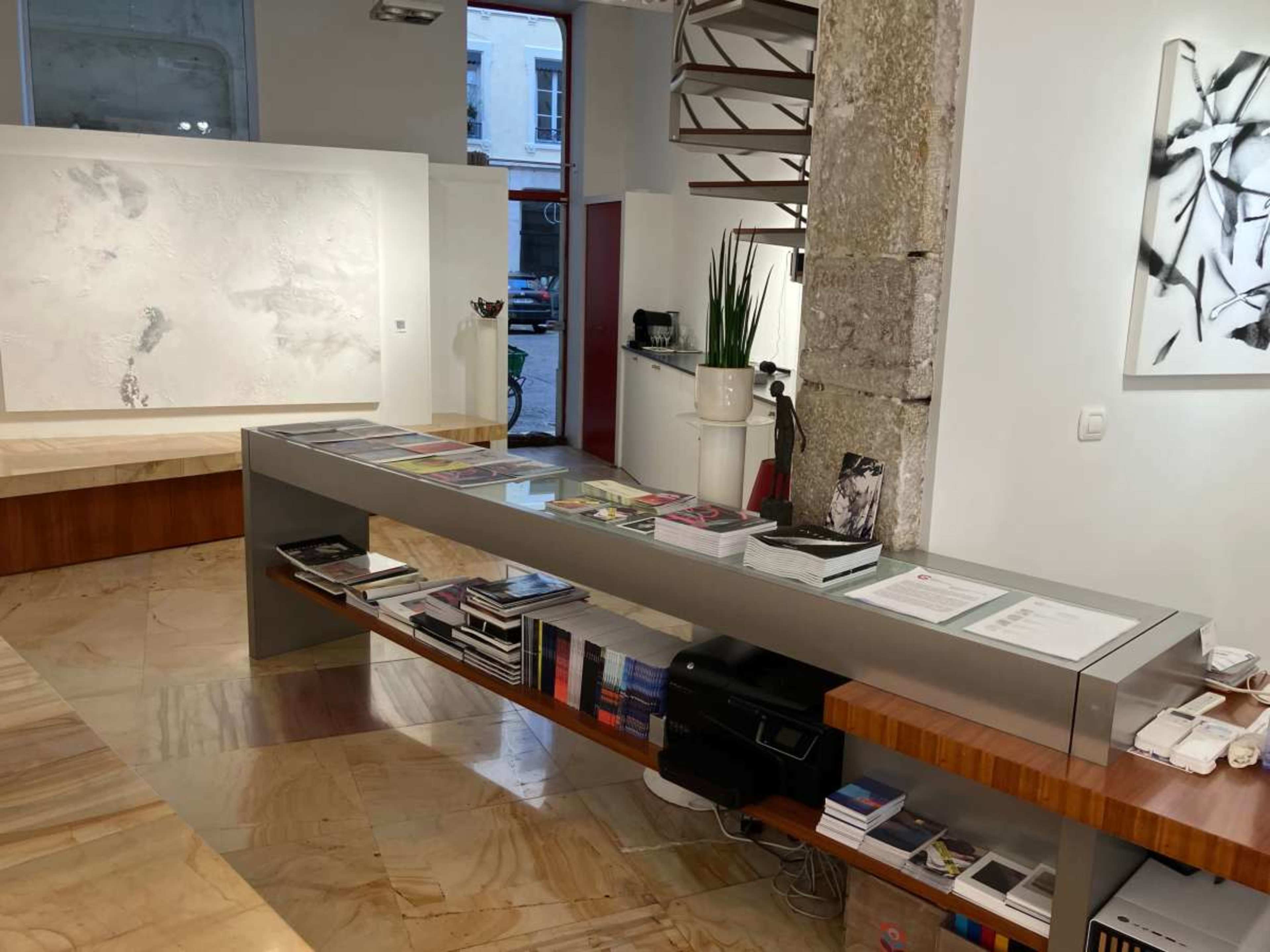The image shows a modern art gallery with a sleek display table featuring various books and catalogs, set against a backdrop of minimalist decor and large windows.