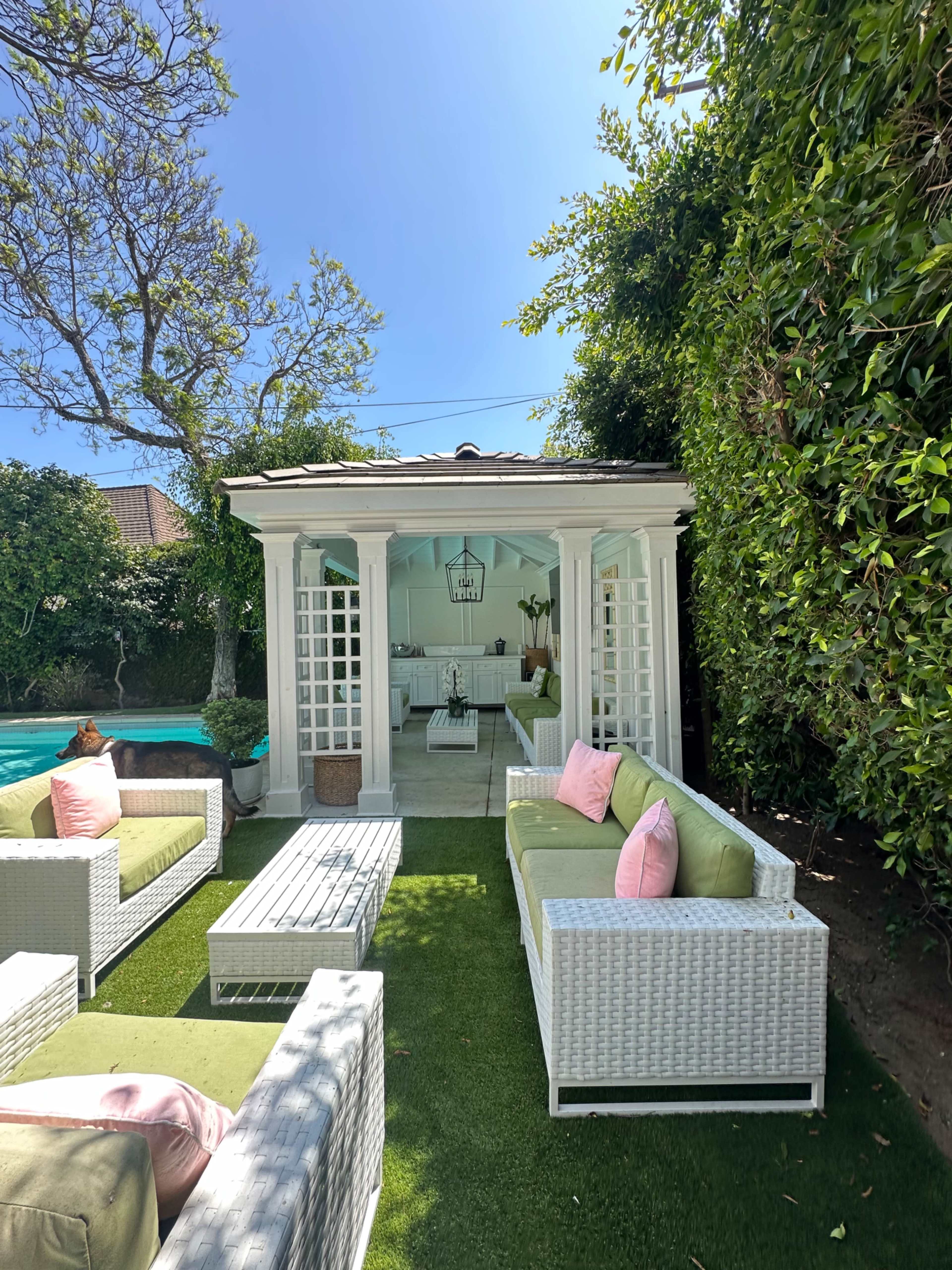 A cozy outdoor lounge area features white wicker furniture with green cushions, surrounding a glass coffee table on a grassy patch next to a poolside cabana.