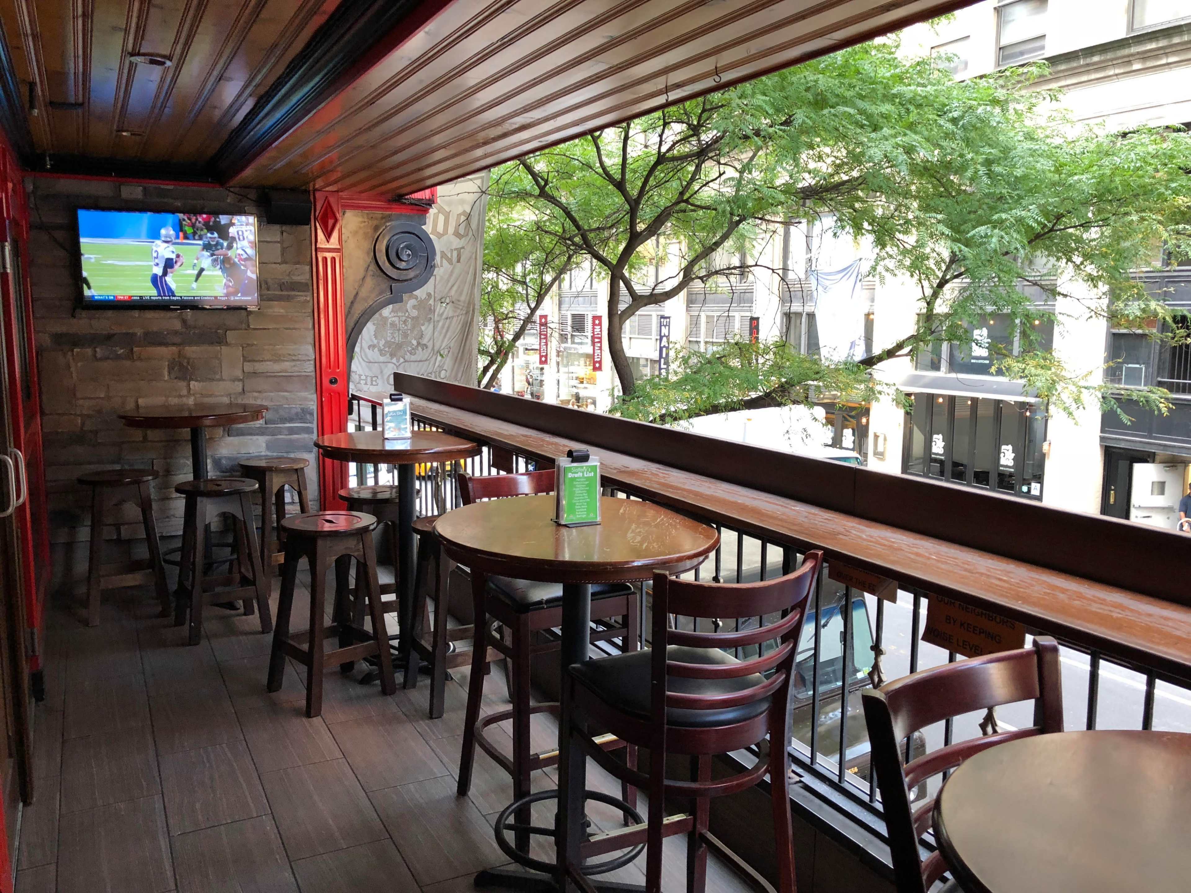 A rooftop outdoor seating area of a bar with wooden tables and chairs, a small TV mounted on the wall, and views of city buildings and trees.