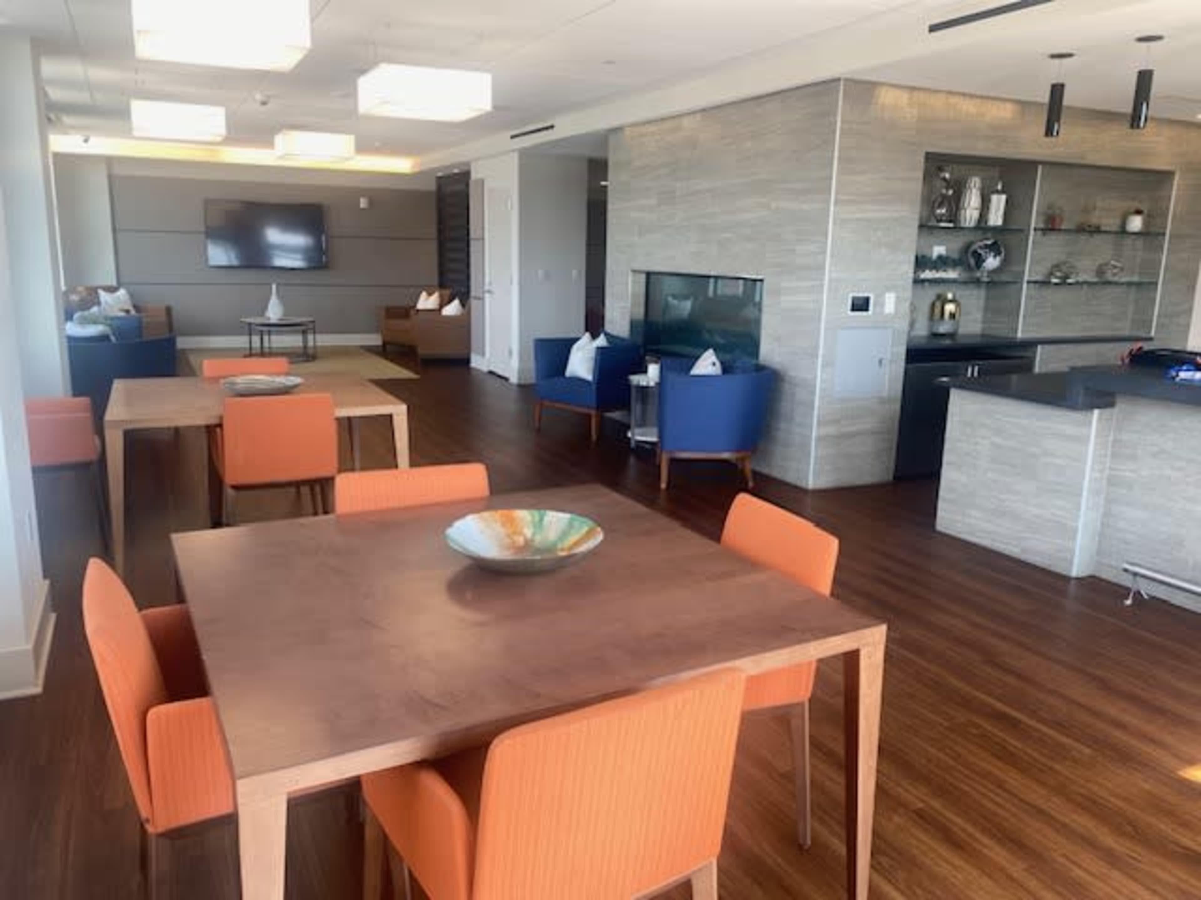 A modern, open living space with wooden tables and orange chairs arranged around a kitchen area and a comfortable seating area.