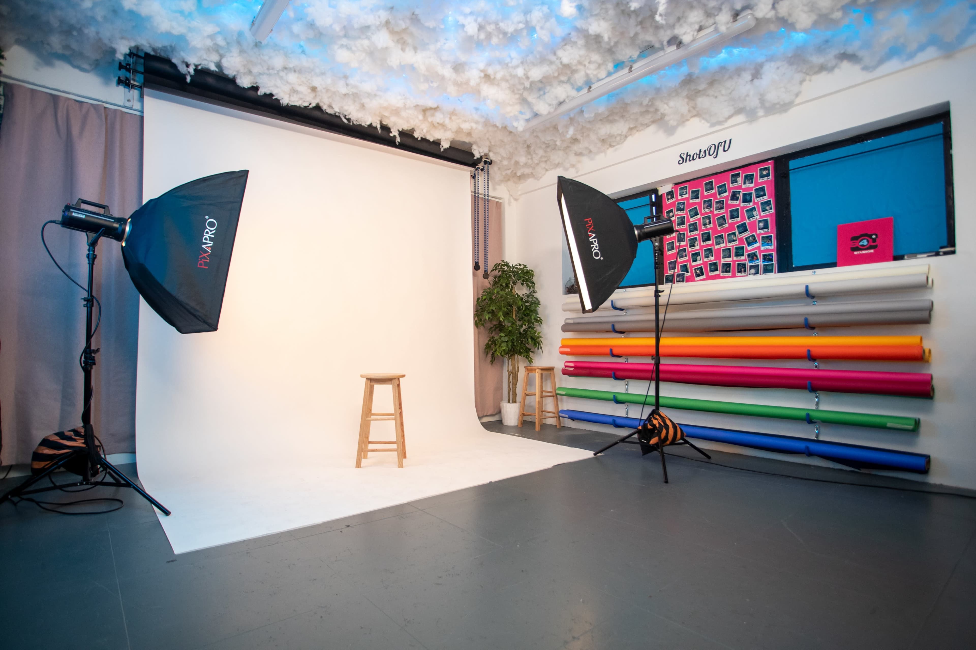 A photography studio with a blank backdrop, two softbox lights, a stool, and a variety of colored backdrop rolls.