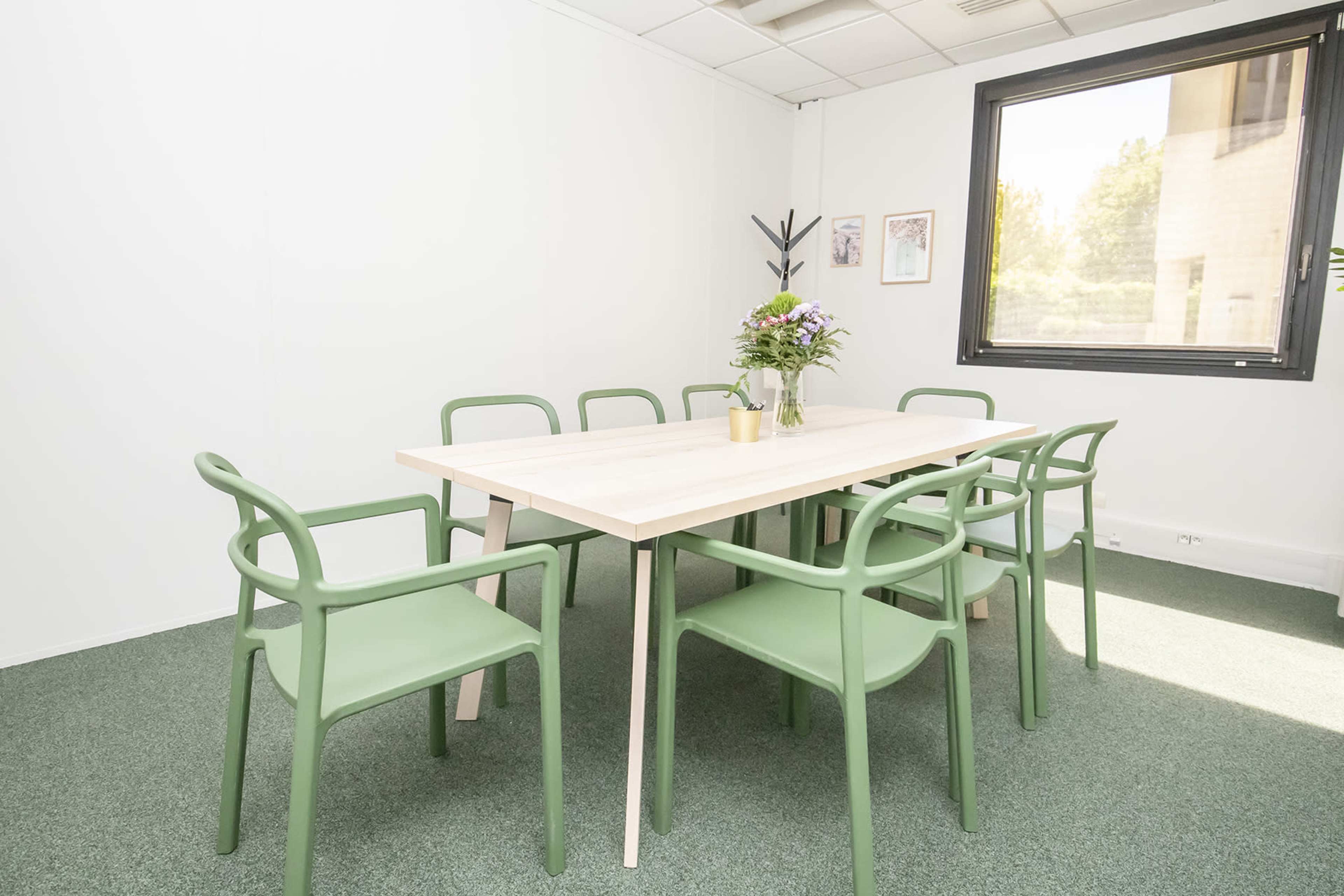 A bright meeting room features a light wooden table surrounded by six green chairs and a vase of flowers.