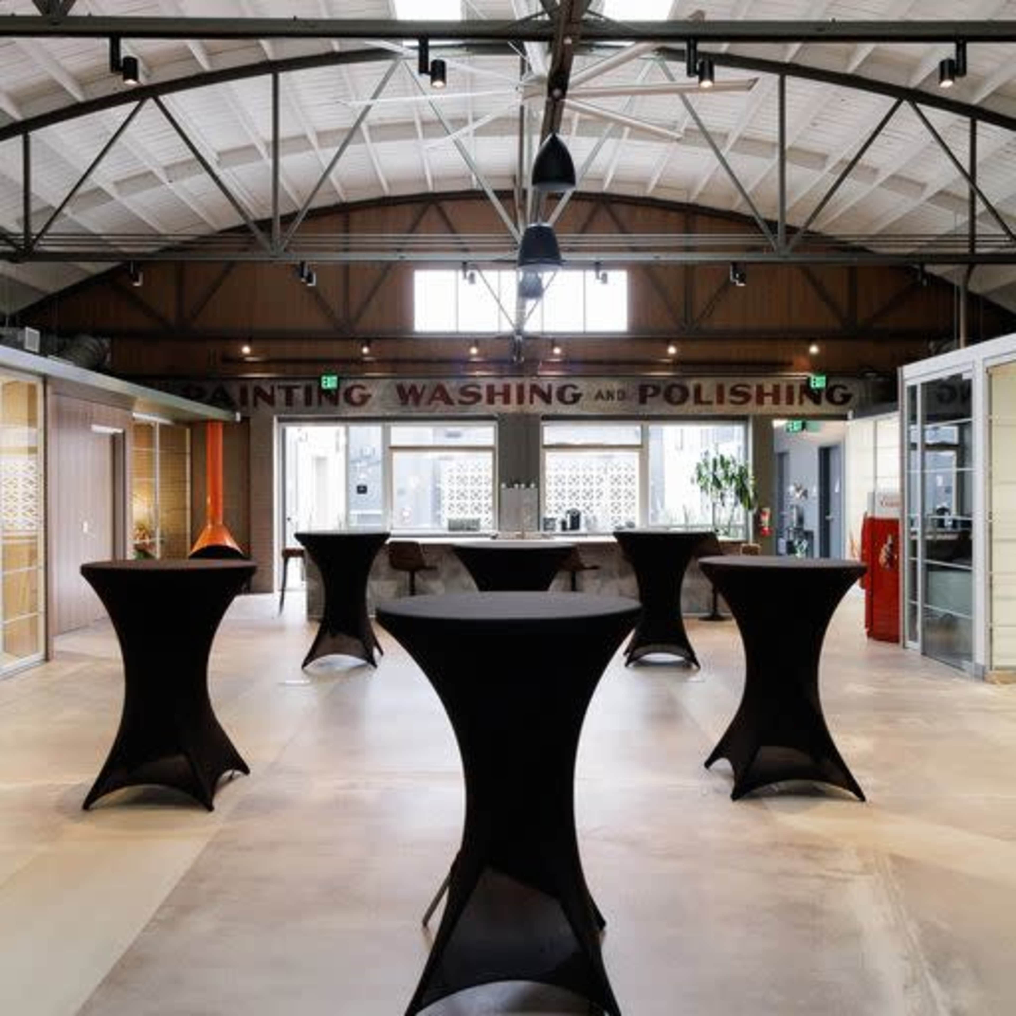 An indoor space with a high ceiling, featuring several tall tables and a central counter area.