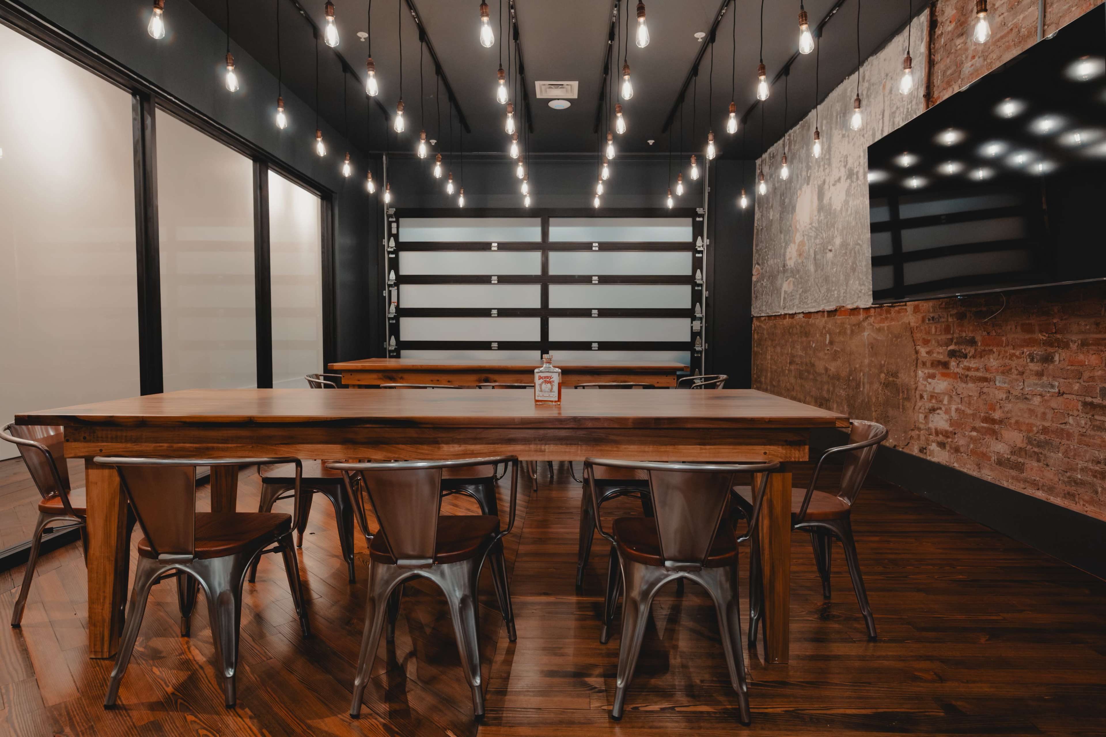 A modern conference room features a long wooden table surrounded by metal chairs, with exposed light bulbs hanging from the ceiling and brick walls.