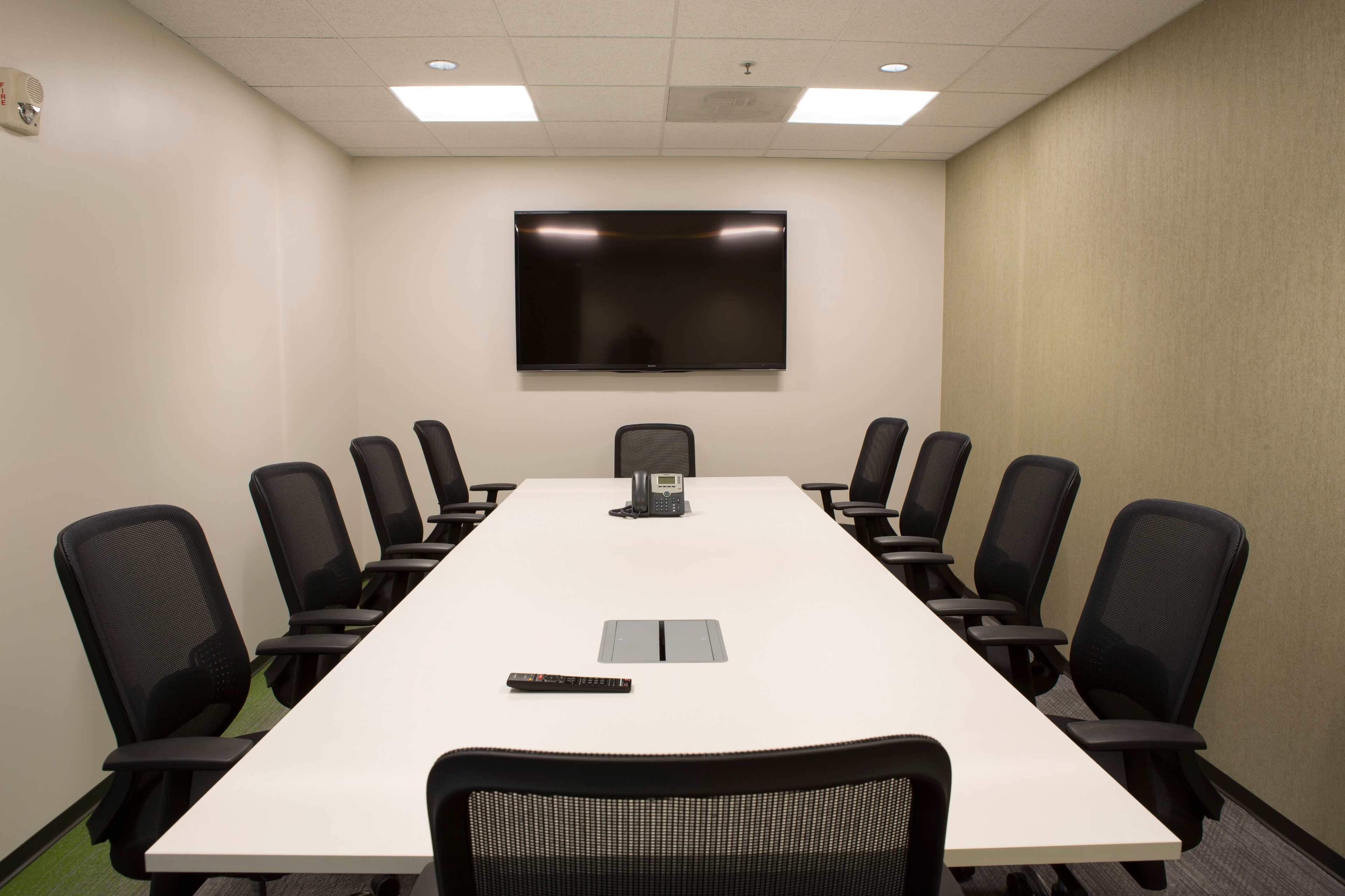 A modern conference room features a long white table surrounded by eight black mesh chairs, with a flat-screen TV mounted on the wall and a phone on the table.