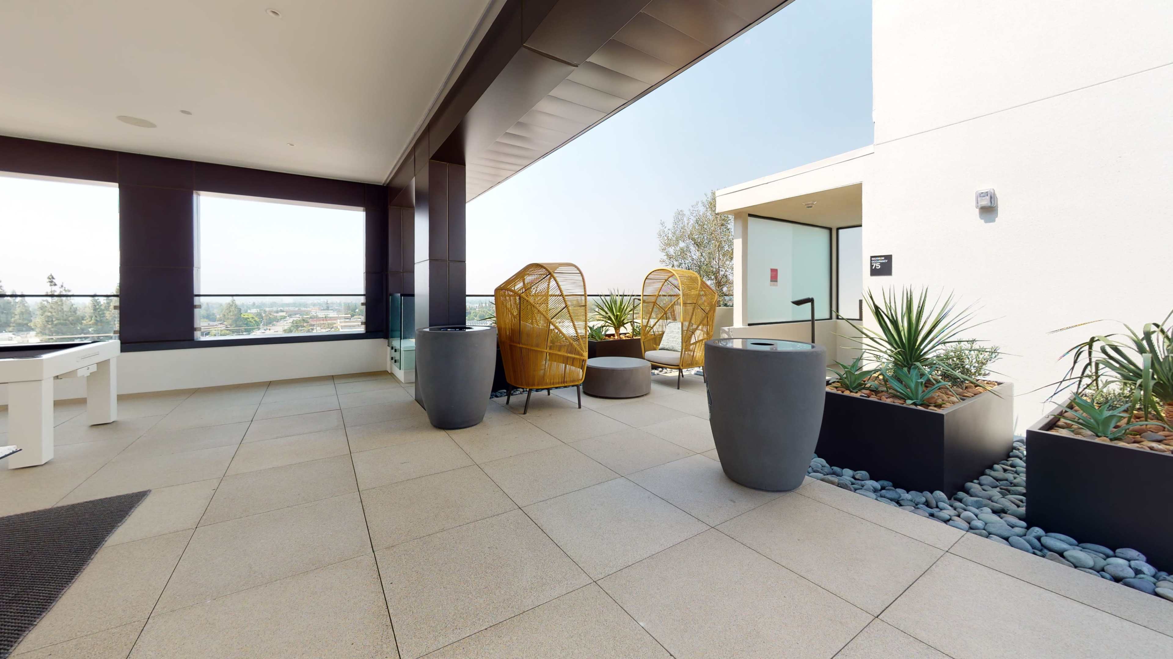 The scene shows a modern balcony area with two yellow wicker chairs, planters, and a view of the horizon.