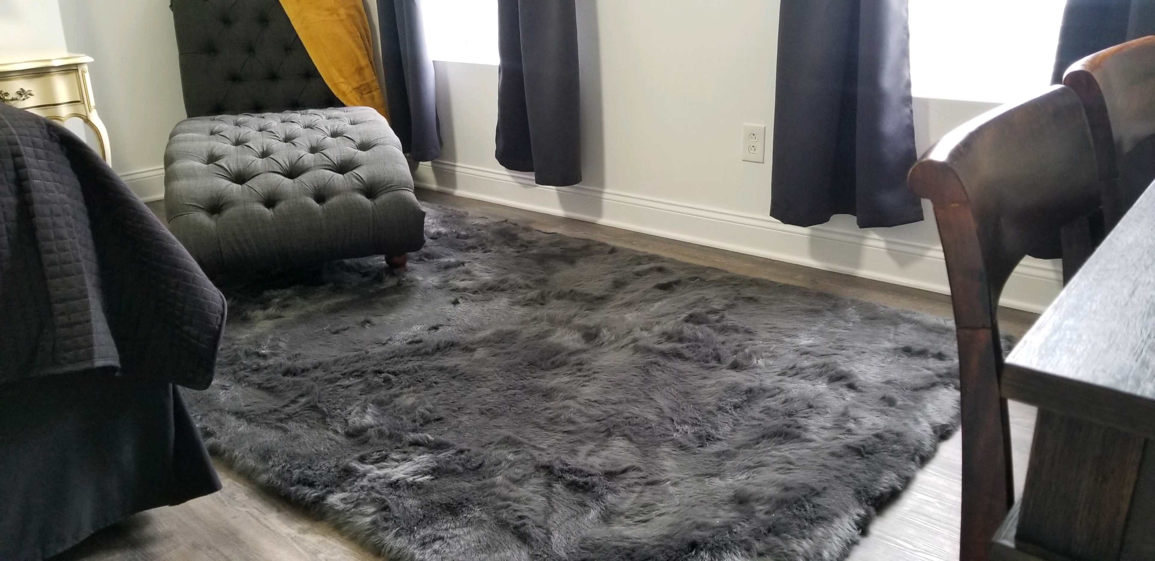 A gray, plush area rug on a hardwood floor, positioned in front of a tufted gray chair and partially covered by a black throw.