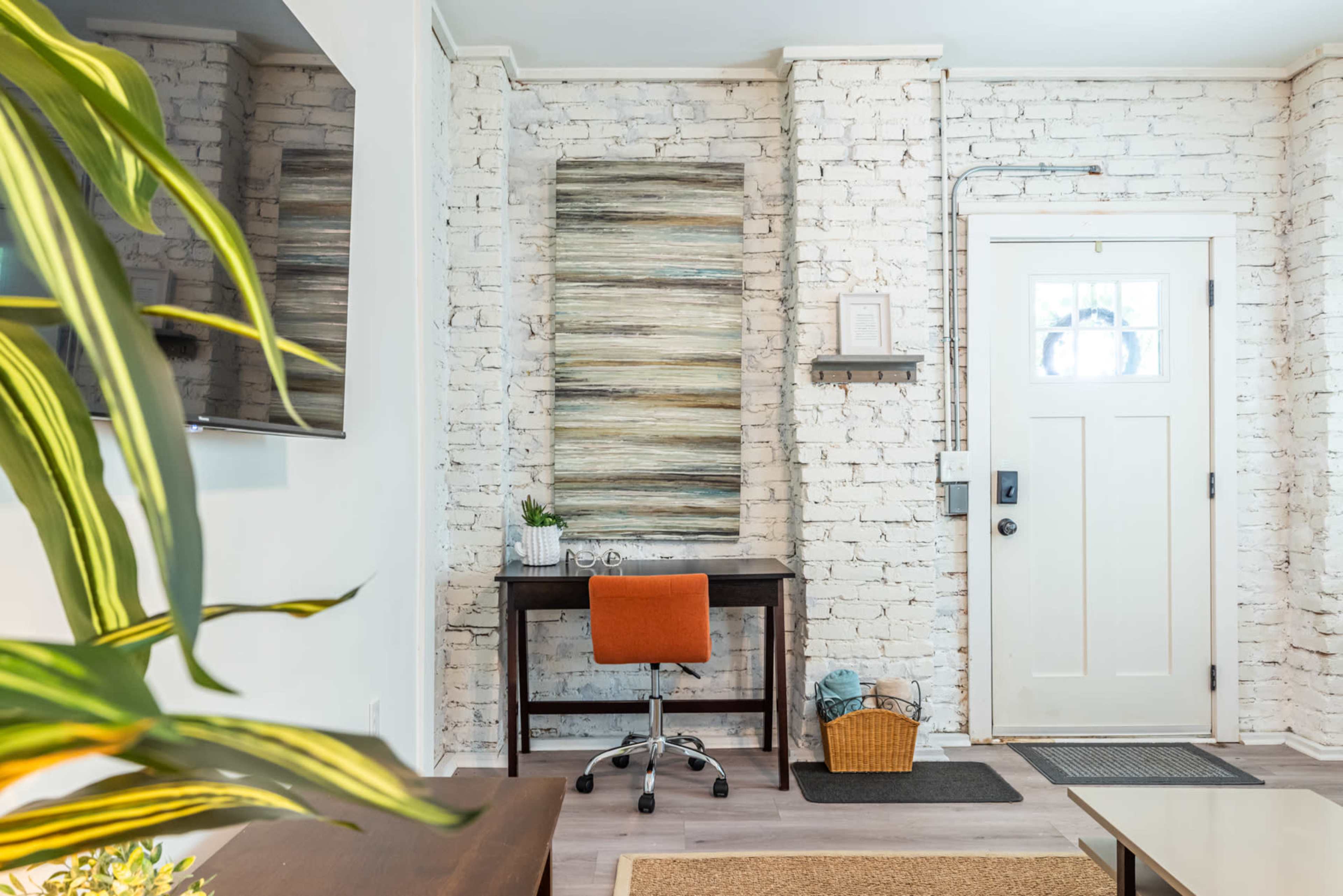 A small workspace with a desk and chair is positioned against a white brick wall, featuring a piece of artwork and a door leading outside.