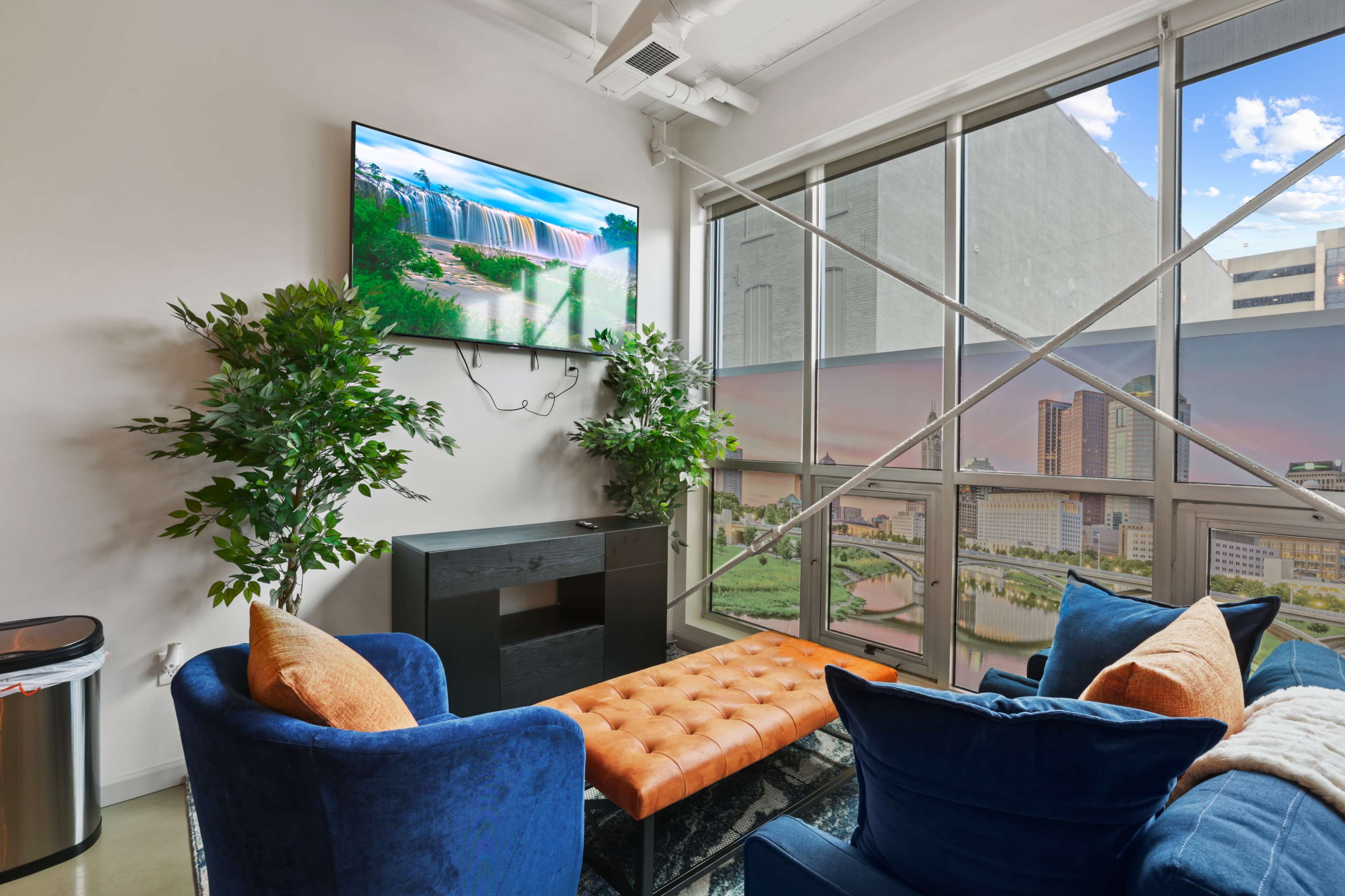 A modern living space with a large TV mounted on the wall, a black media console, two blue chairs, and a tufted orange ottoman, all positioned near a large window with a cityscape view.