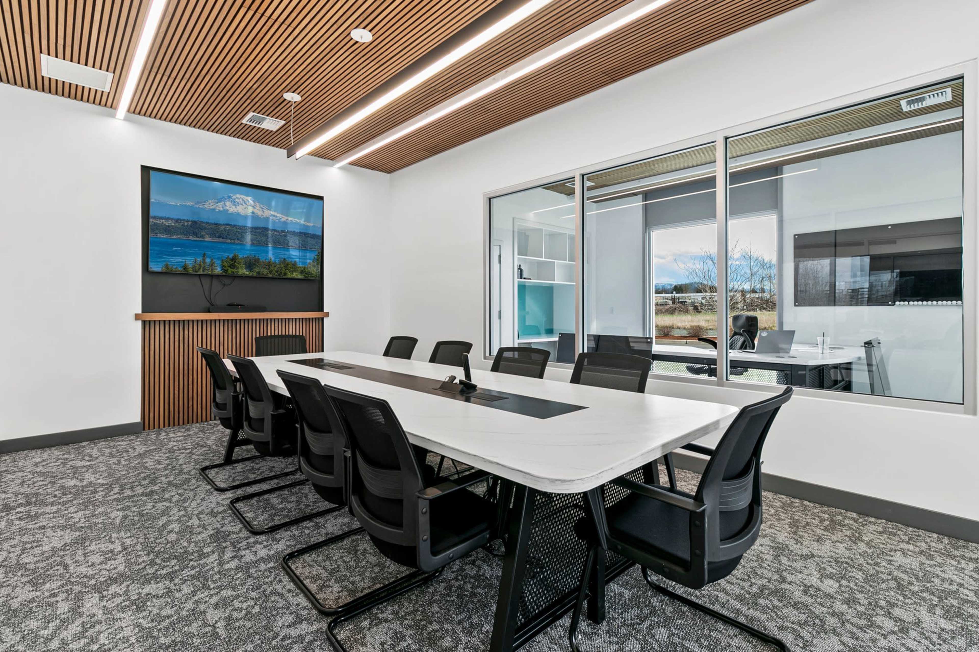 The image shows a modern conference room featuring a long table surrounded by black chairs, a large screen mounted on the wall, and floor-to-ceiling windows providing natural light.