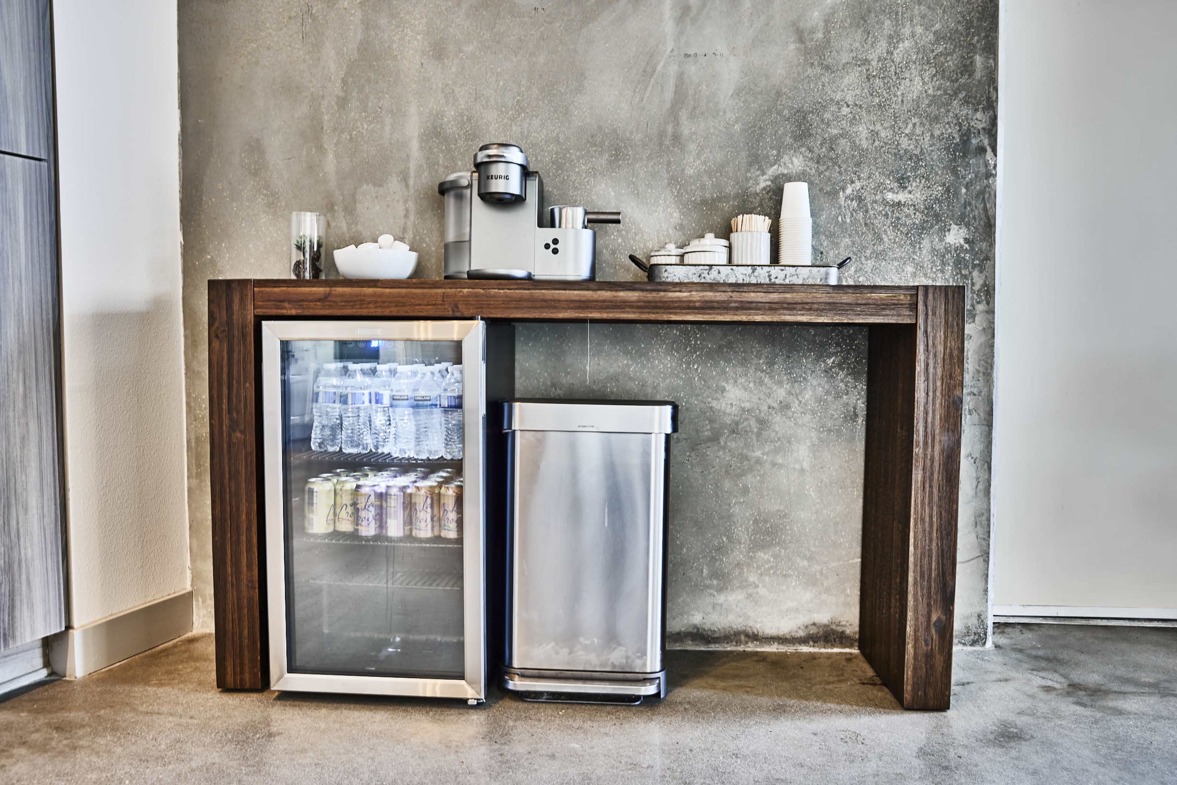 A wooden table holds a mini refrigerator, a coffee maker, and a tray with utensils and cups against a concrete wall.
