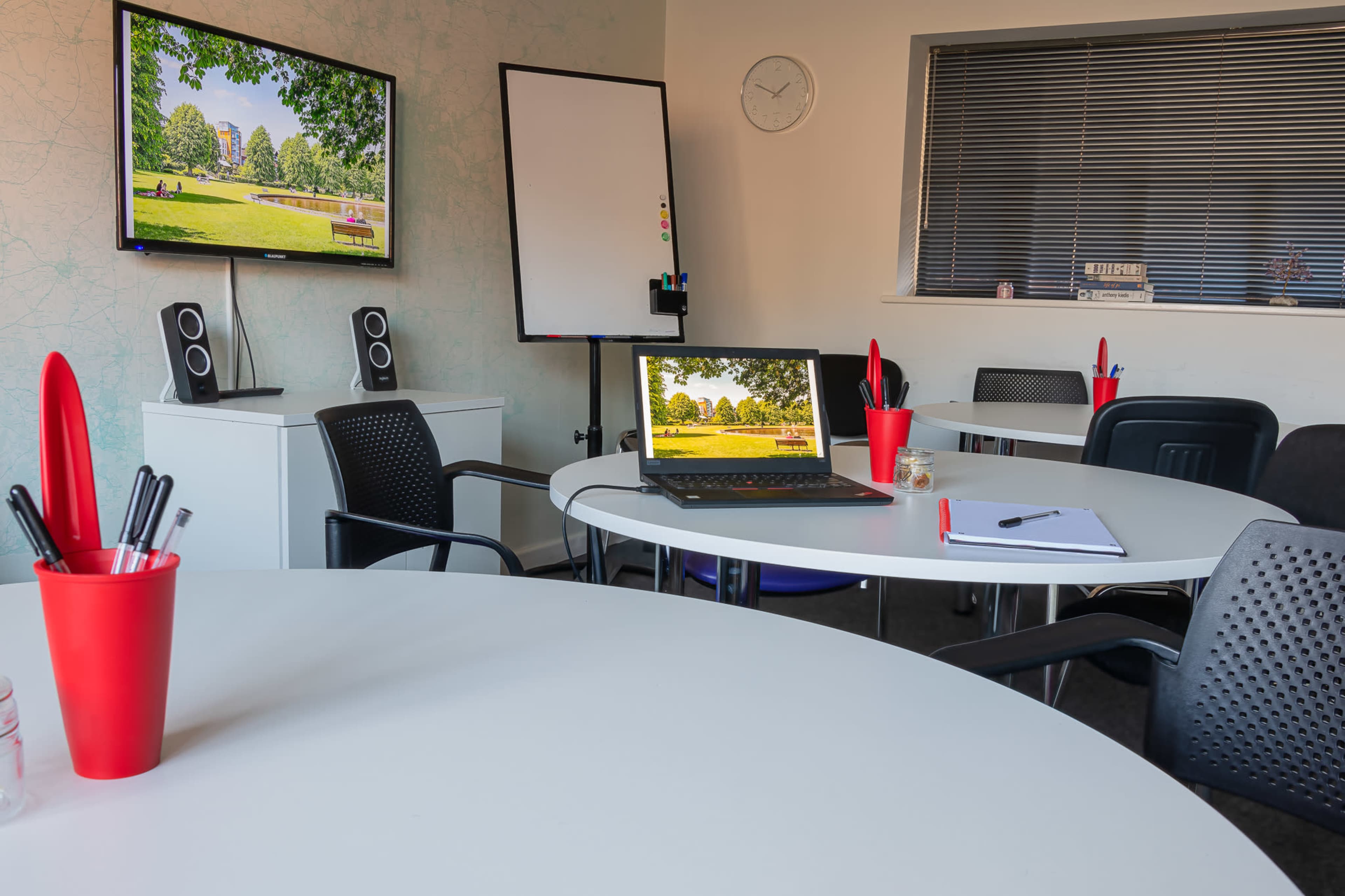 A modern meeting room features a laptop, a whiteboard, and two screens displaying a park scene, with a centerpiece of red pen holders on the tables.