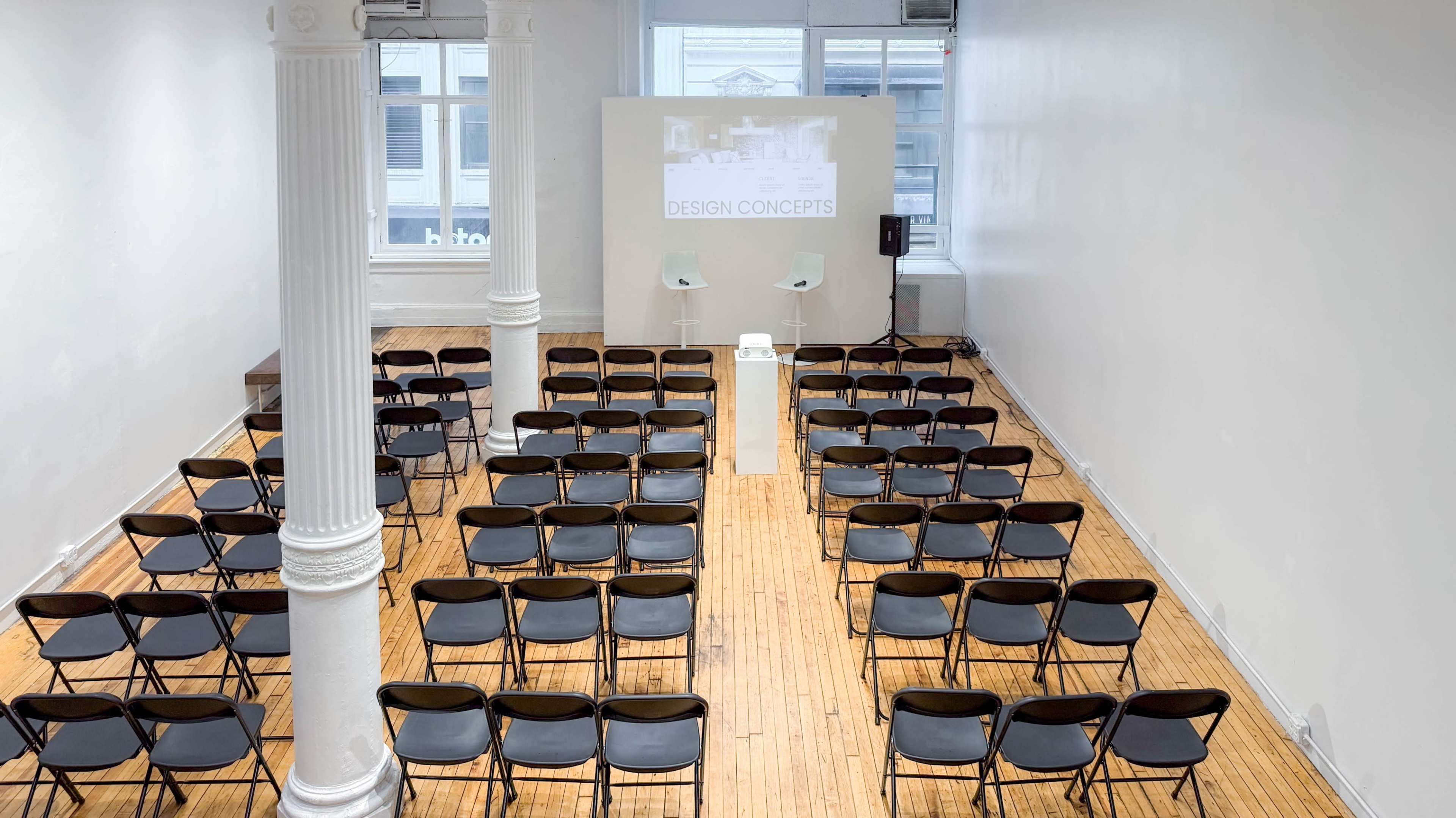 The image shows a spacious, empty presentation room with rows of black chairs arranged in front of a white wall, where a projection screen displays "DESIGN CONCEPTS."