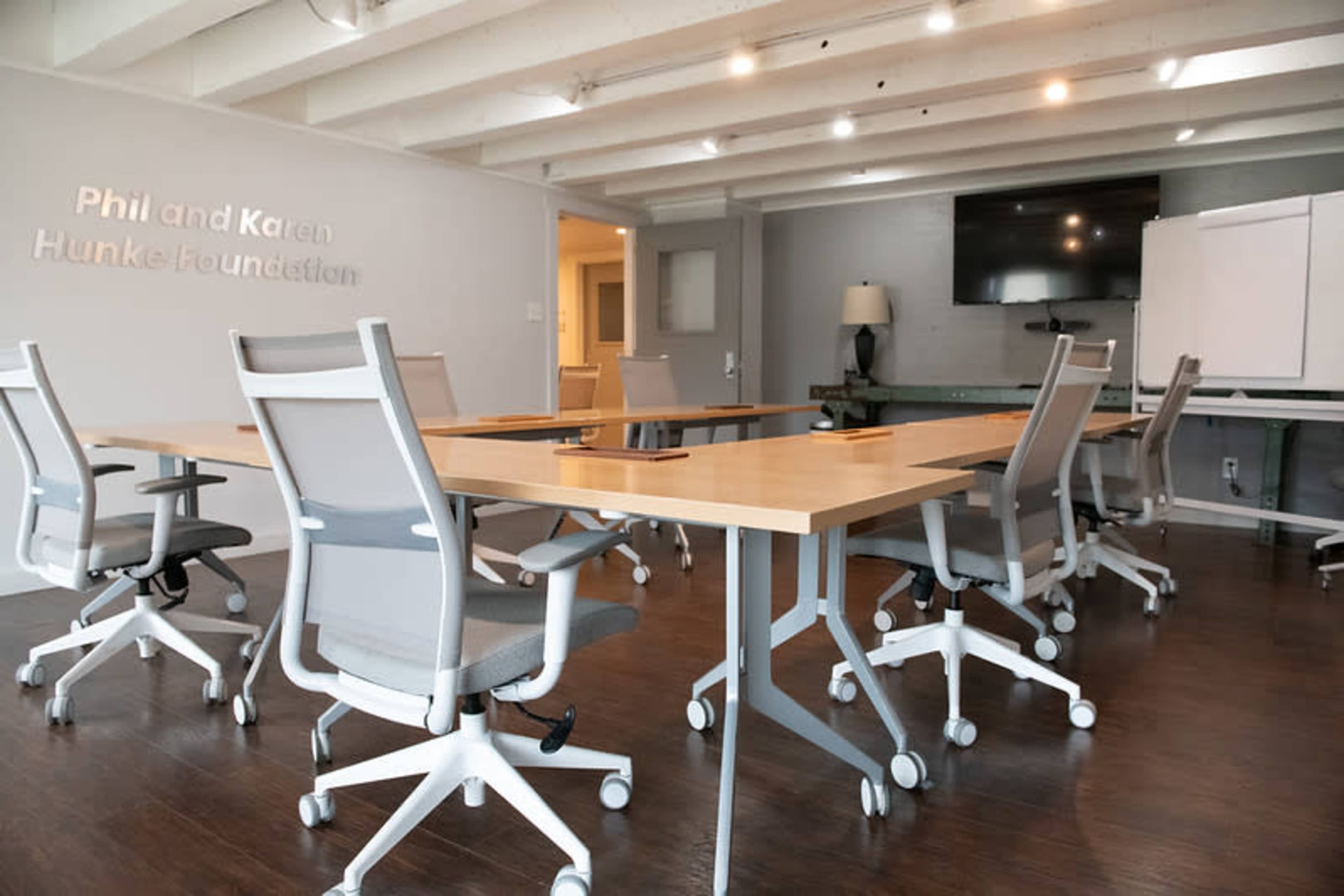 The image shows a spacious conference room with several light-colored tables and ergonomic chairs arranged around them, featuring a large television screen on the wall and a whiteboard in the background.