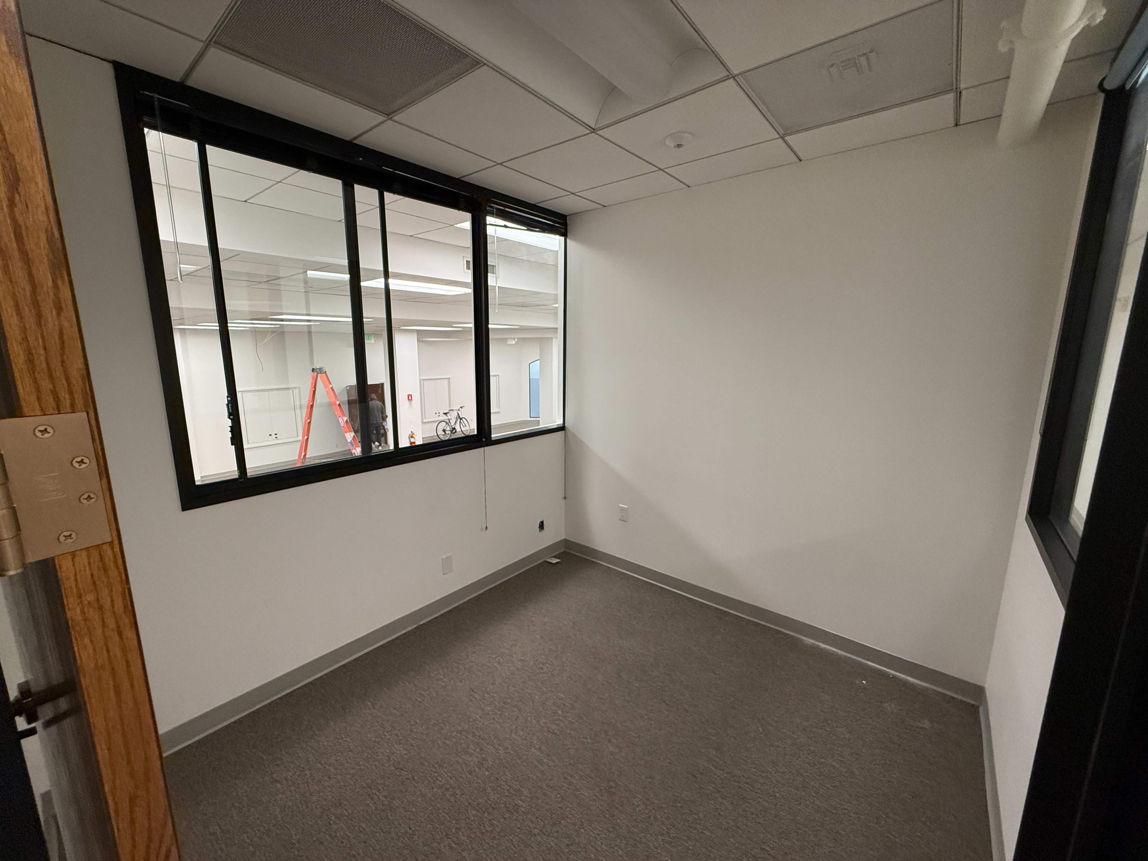 The image shows a vacant office space with gray carpet, white walls, and a large window overlooking a hallway with a ladder and bicycles visible.