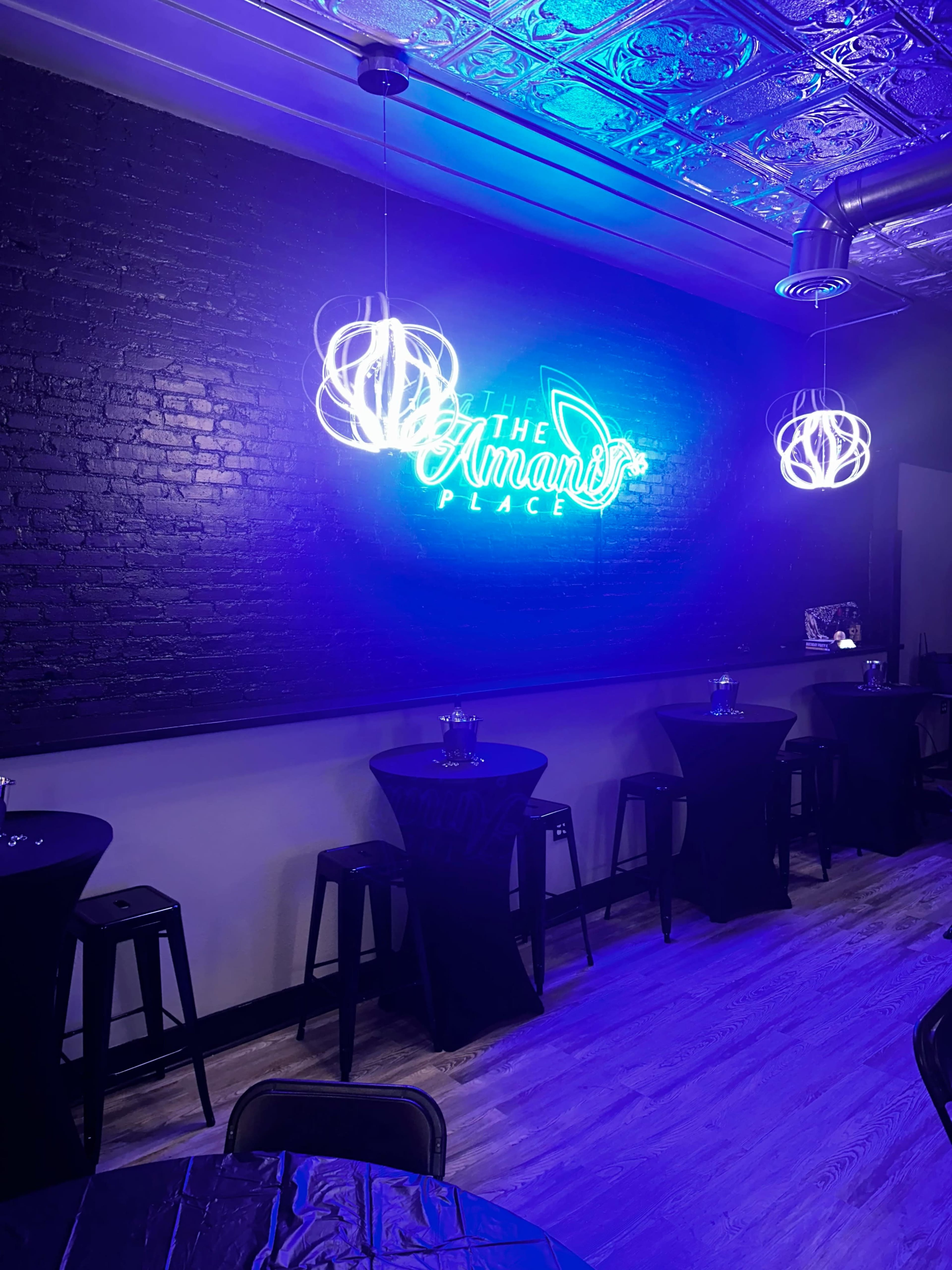 The image shows a dimly lit event space with a neon sign reading "The Amanda's Place" and several black tables covered in tablecloths.