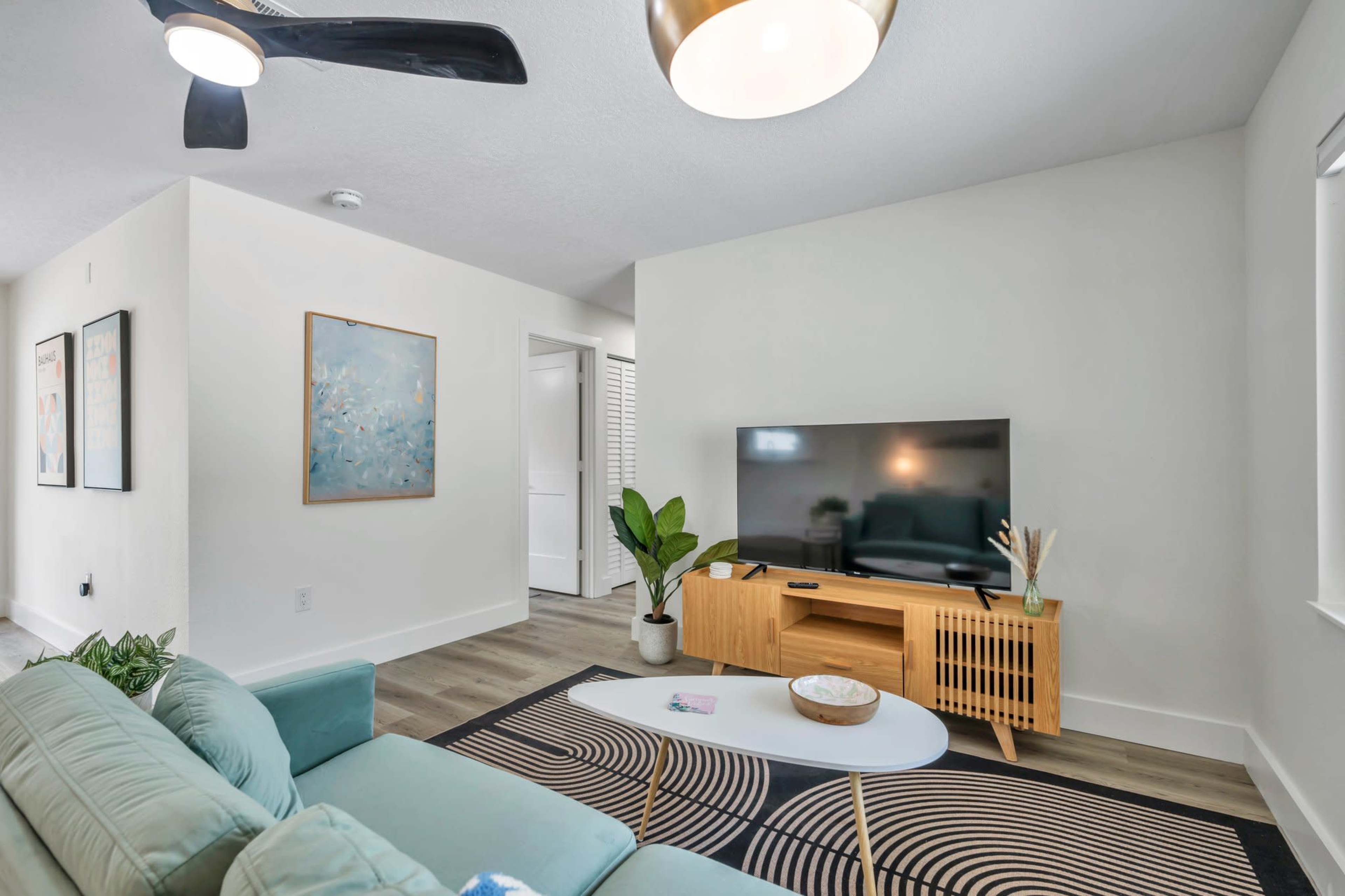 The living room features a teal sofa, a wooden TV stand, a large television, and a decorative plant against a light-colored wall.