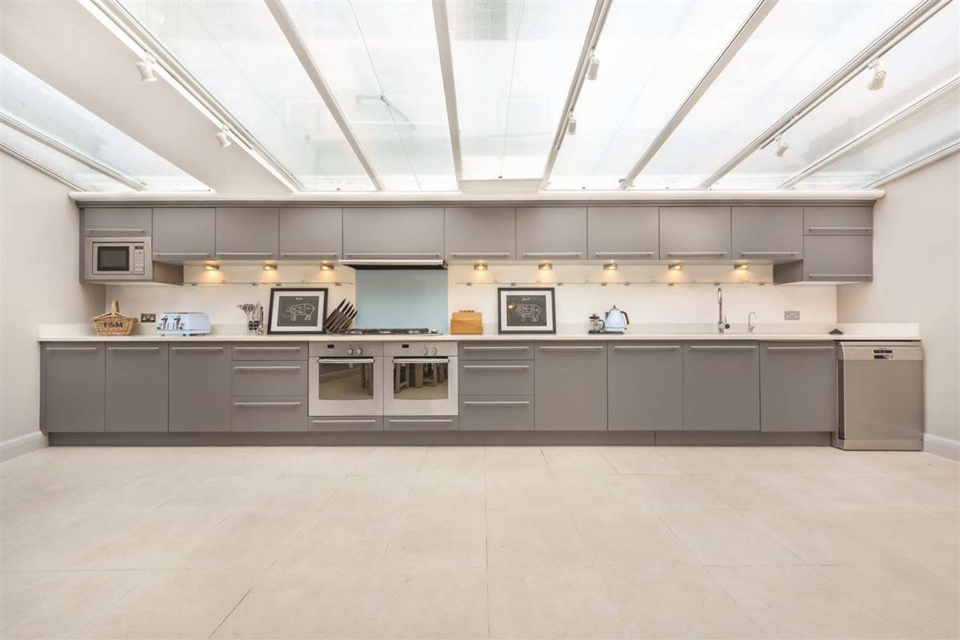 A modern kitchen features a long, gray cabinetry setup with a central cooking area and ample counter space, illuminated by under-cabinet lighting and a glass roof.