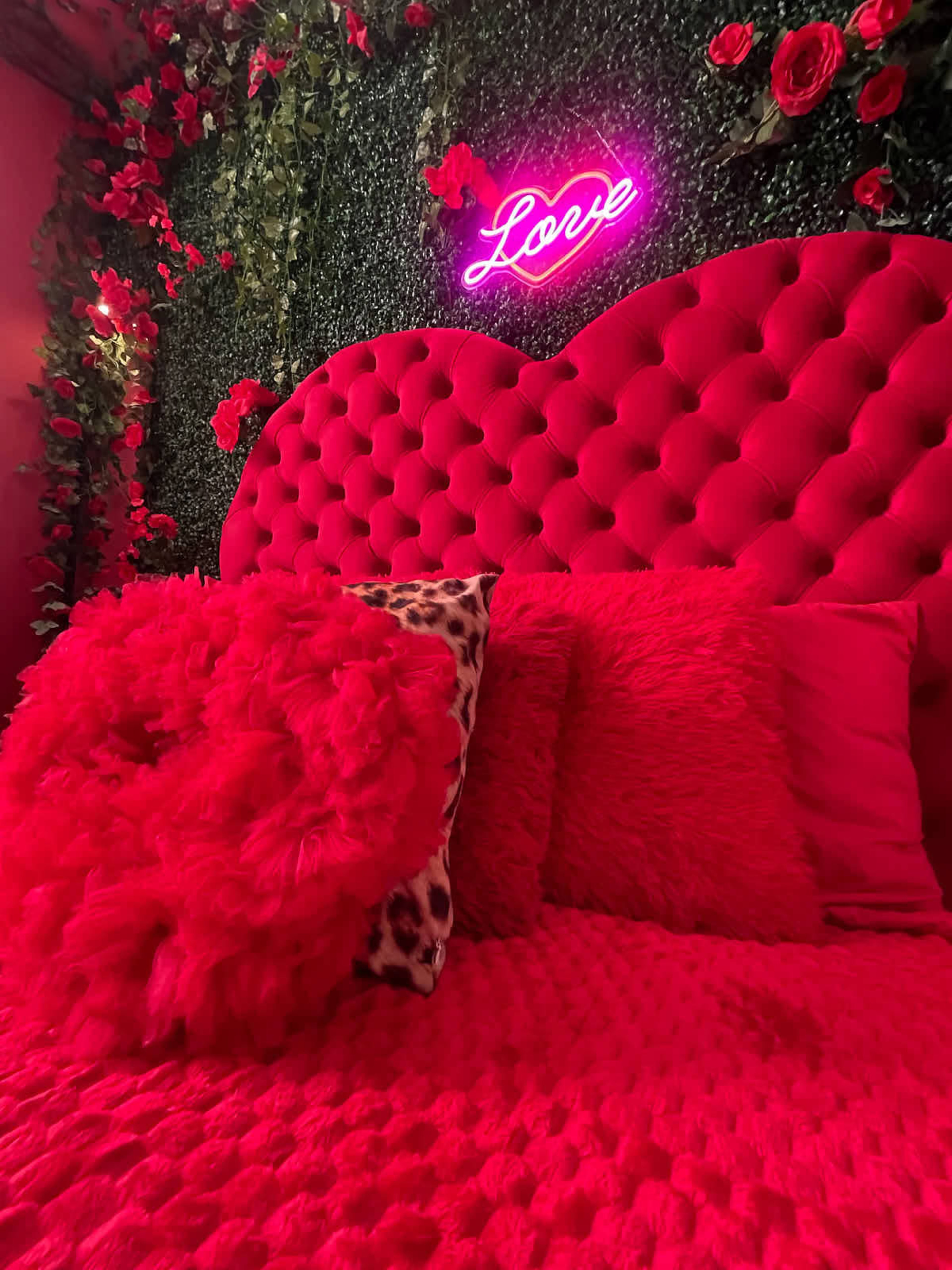 A vibrant bedroom features a pink tufted headboard, red pillows, and a neon sign that reads "Love," surrounded by artificial greenery and floral decor.