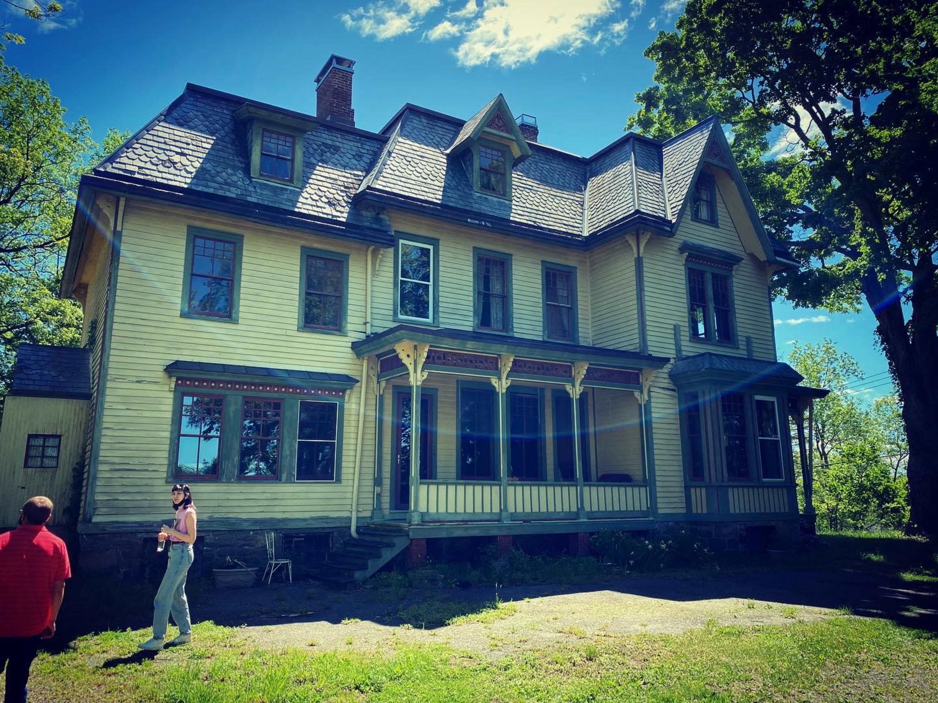 A large, two-story yellow Victorian-style house with multiple gables and a covered porch stands under a clear blue sky.