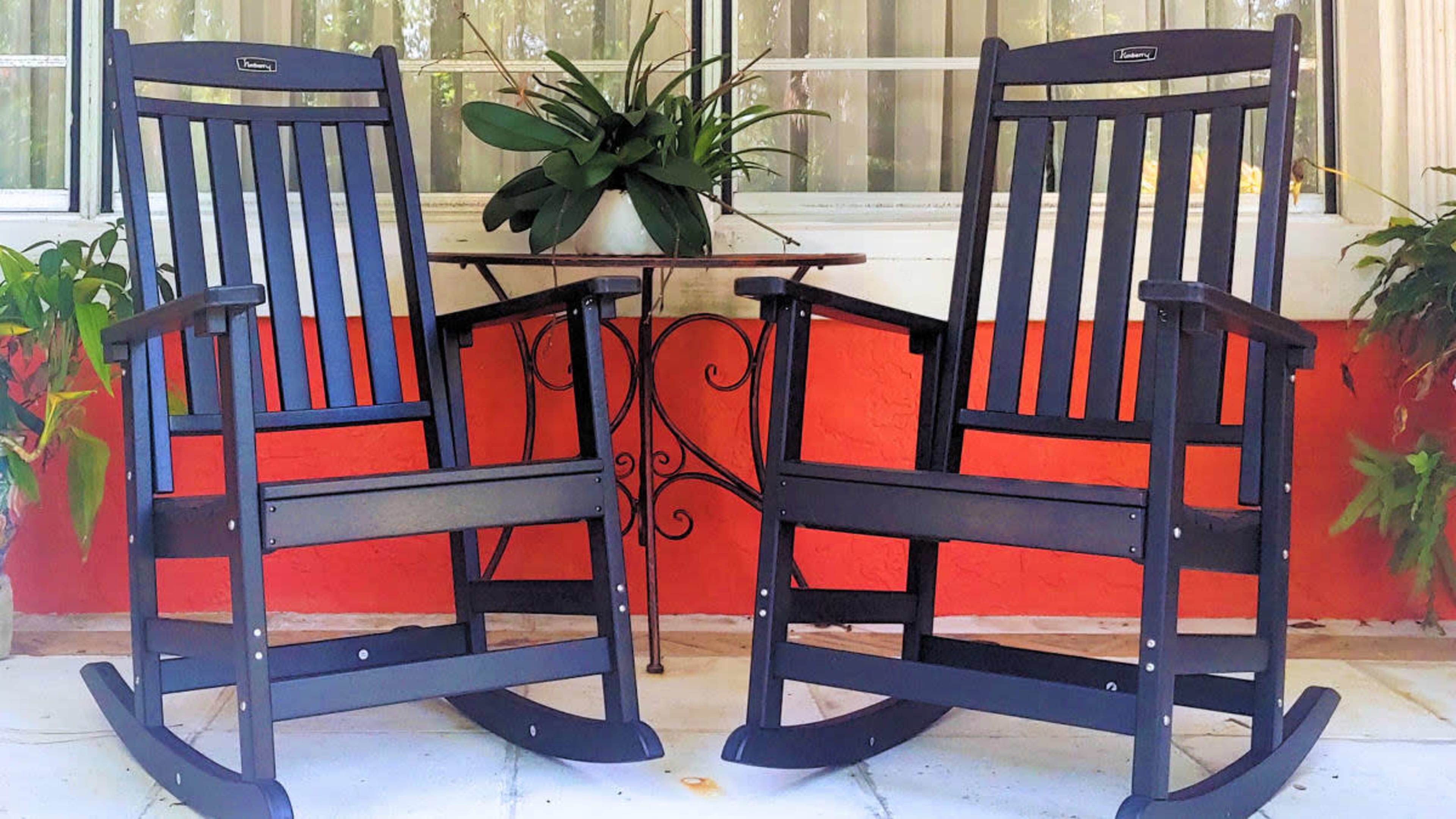 Two black rocking chairs are positioned in front of a window, with a small table between them and potted plants nearby.