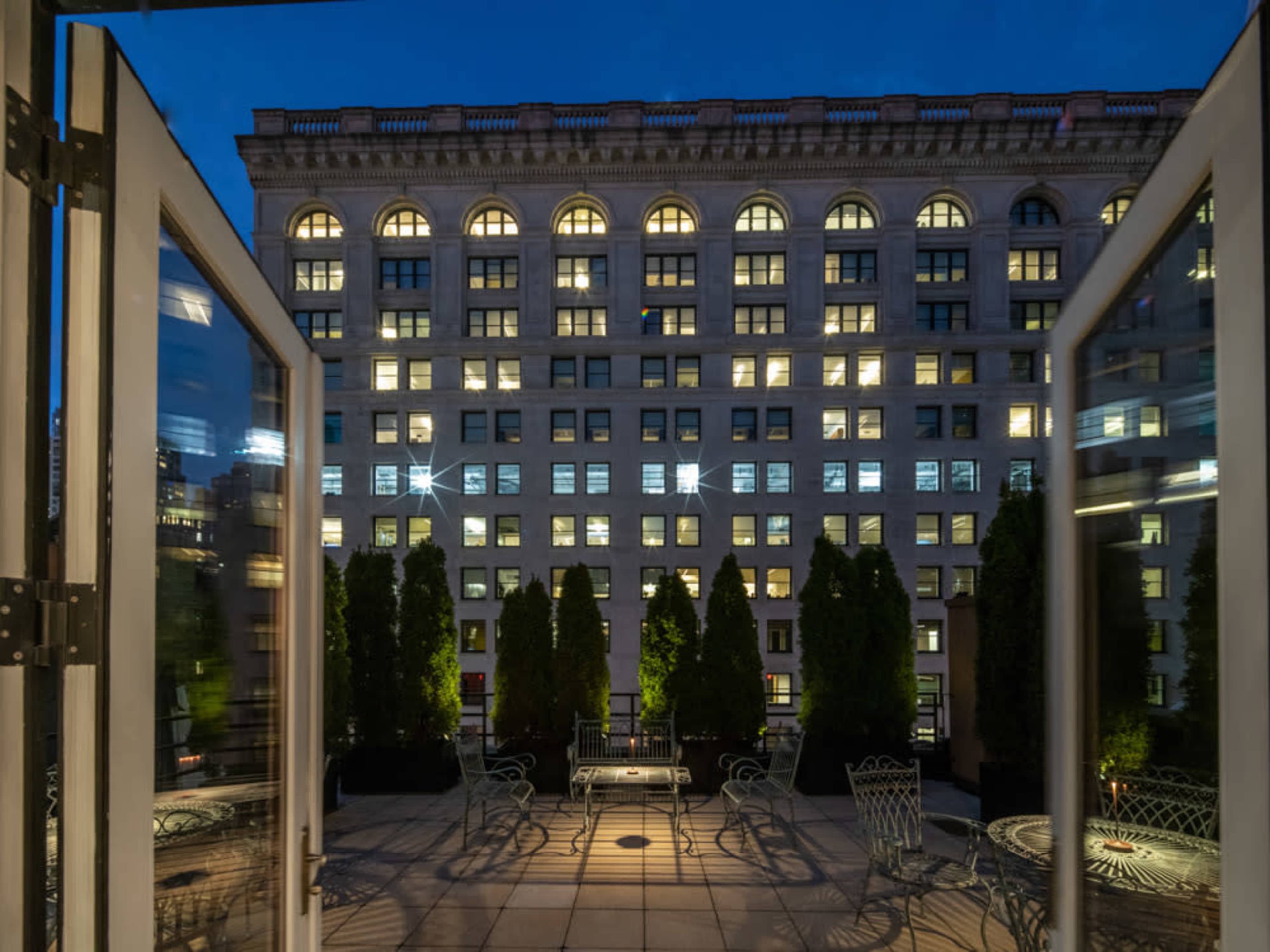 The image shows an open balcony with iron furniture overlooking a building facade illuminated at night, featuring rows of windows.