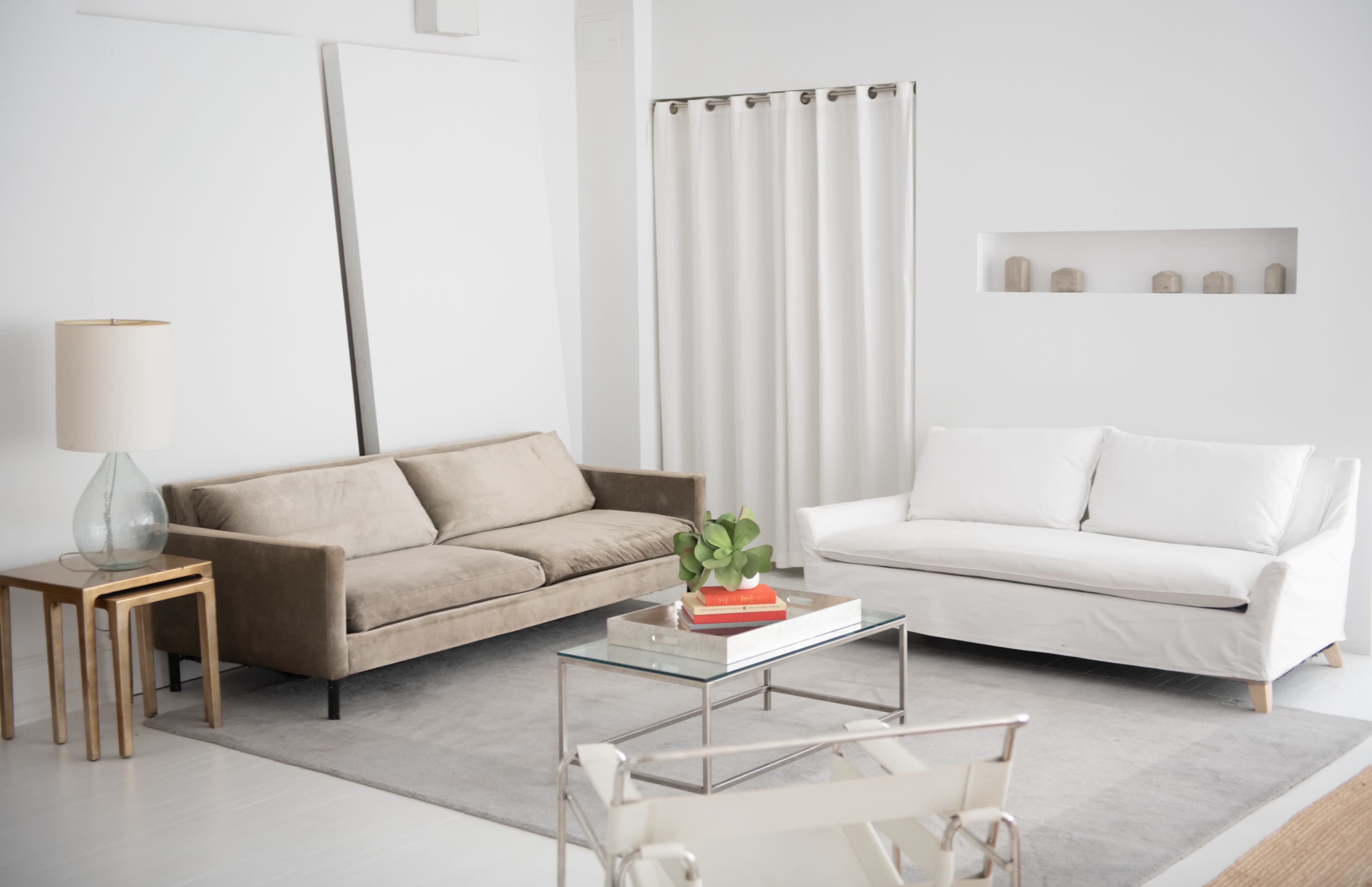 A modern living room with a beige sofa, a white couch, a glass coffee table, and a light-colored rug, all set against white walls.