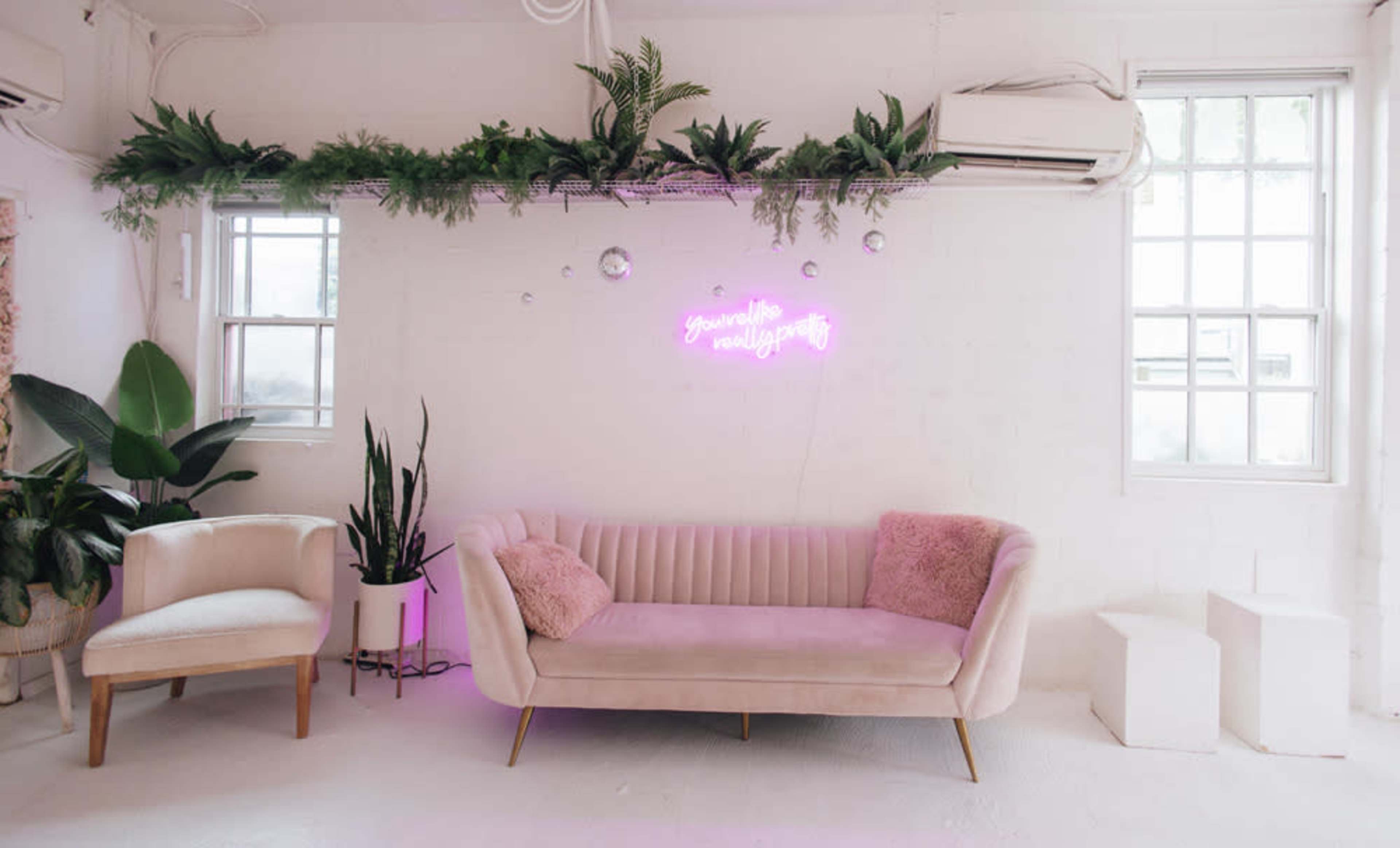 A light-colored room featuring a pink couch with a plush pillow, a small armchair, and greenery displayed above, accompanied by a neon sign that reads "You are enough."