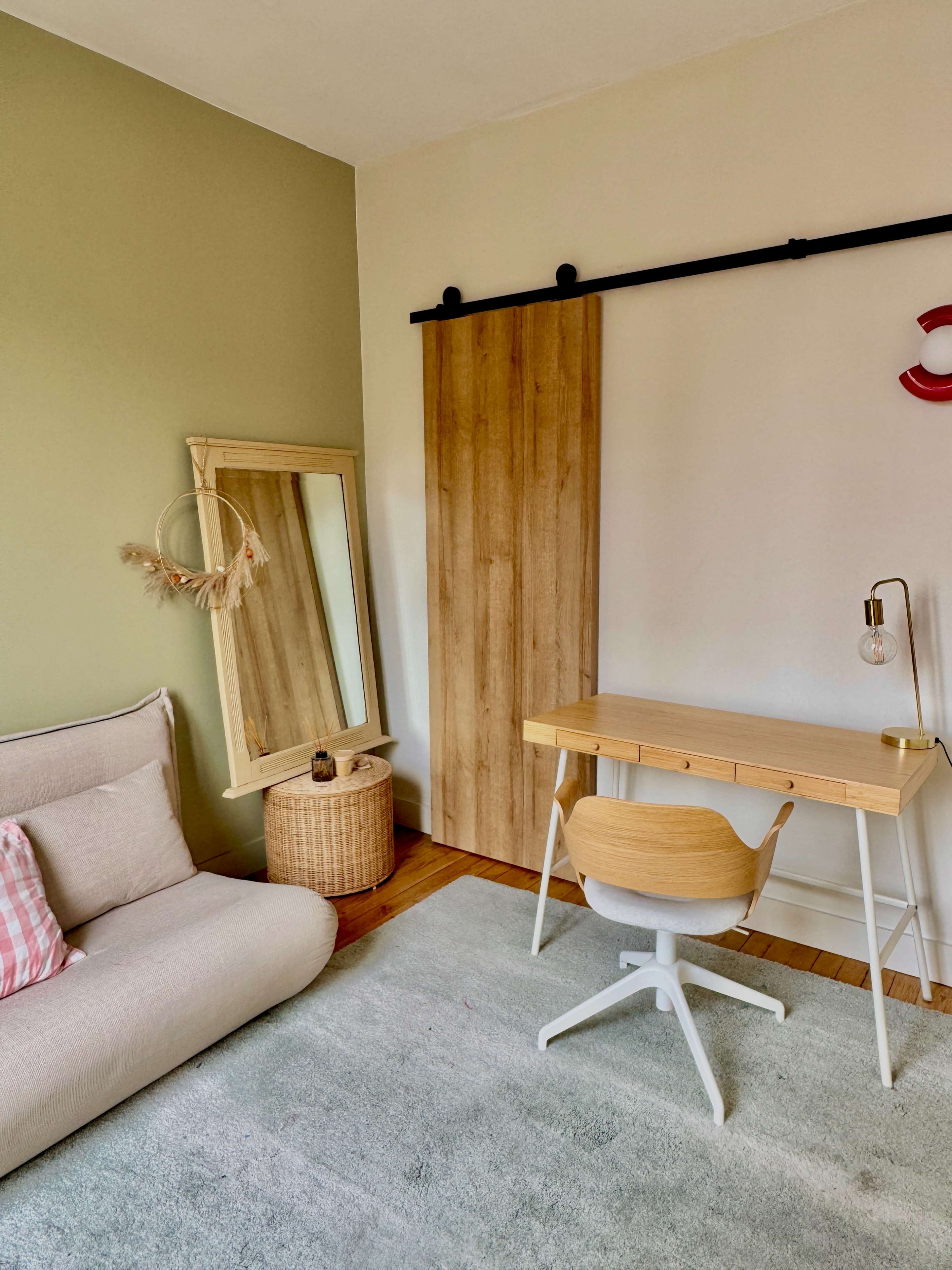 The image shows a light, minimalistic room featuring a wooden desk with a chair, a large mirror, a soft seating area, and a sliding wooden panel against a light green wall.