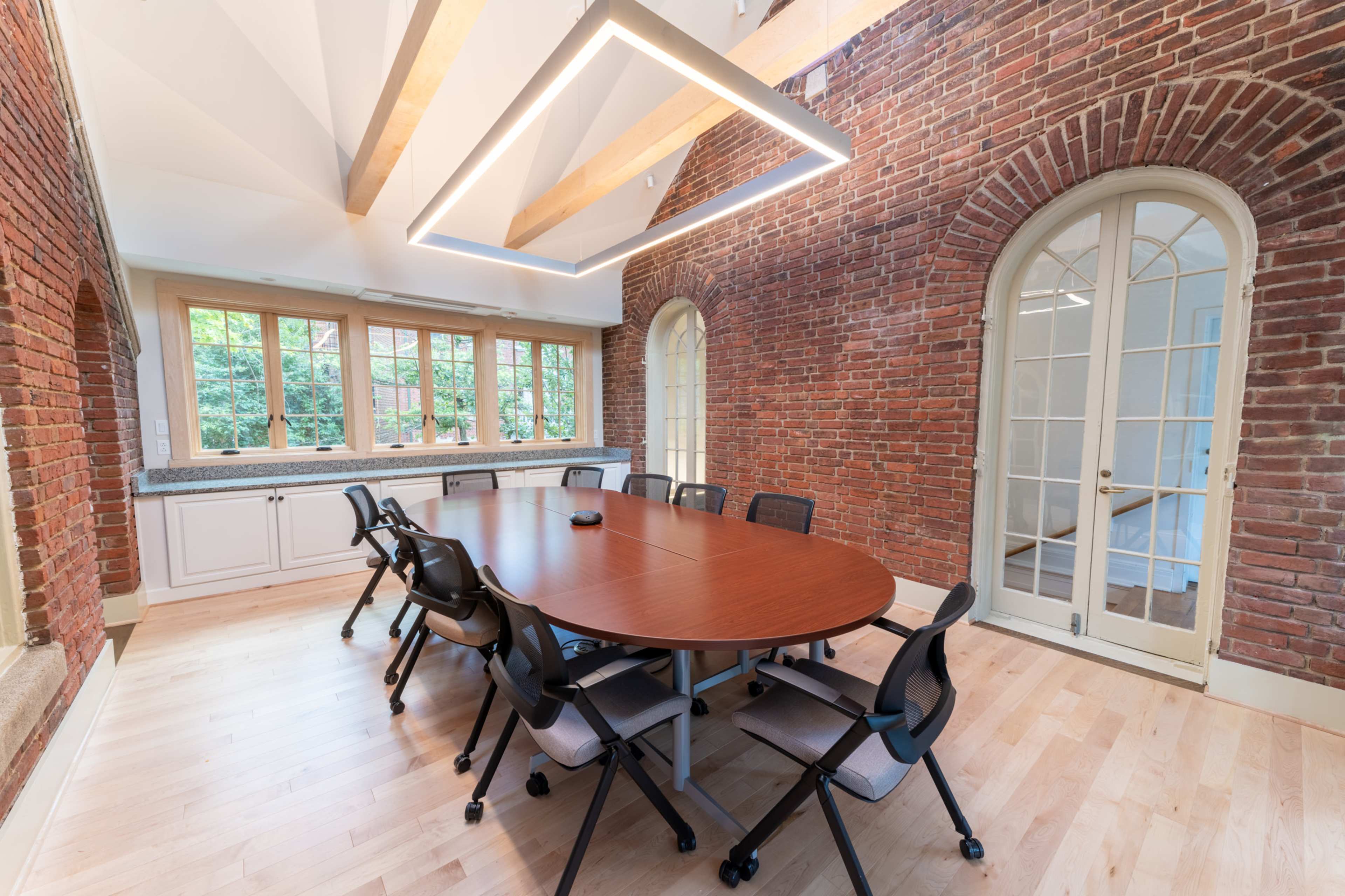 A modern conference room featuring a round wooden table surrounded by black chairs, with exposed brick walls and large windows allowing natural light.
