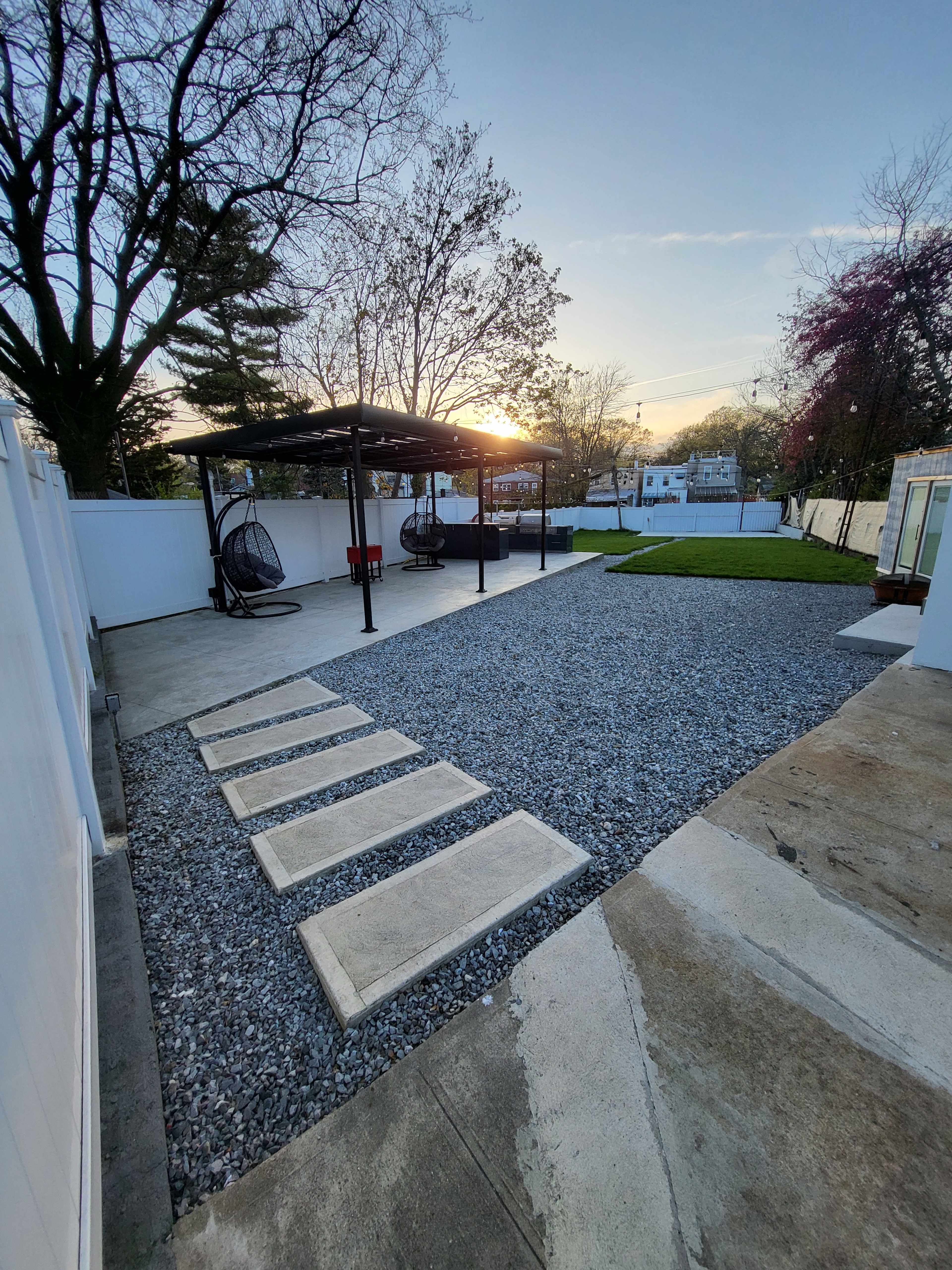 A backyard with a gravel area, concrete stepping stones, a covered seating area, and a green lawn under a sunset sky.