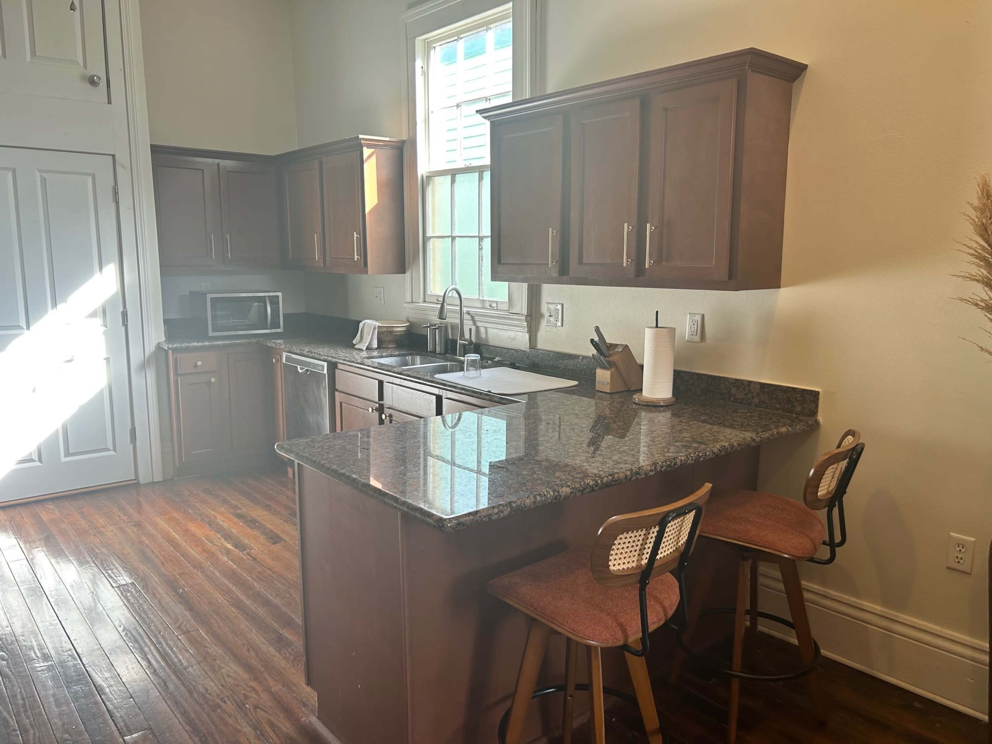 The image shows a well-lit kitchen with wooden cabinets, a granite countertop, a sink, and two barstools at a kitchen island.