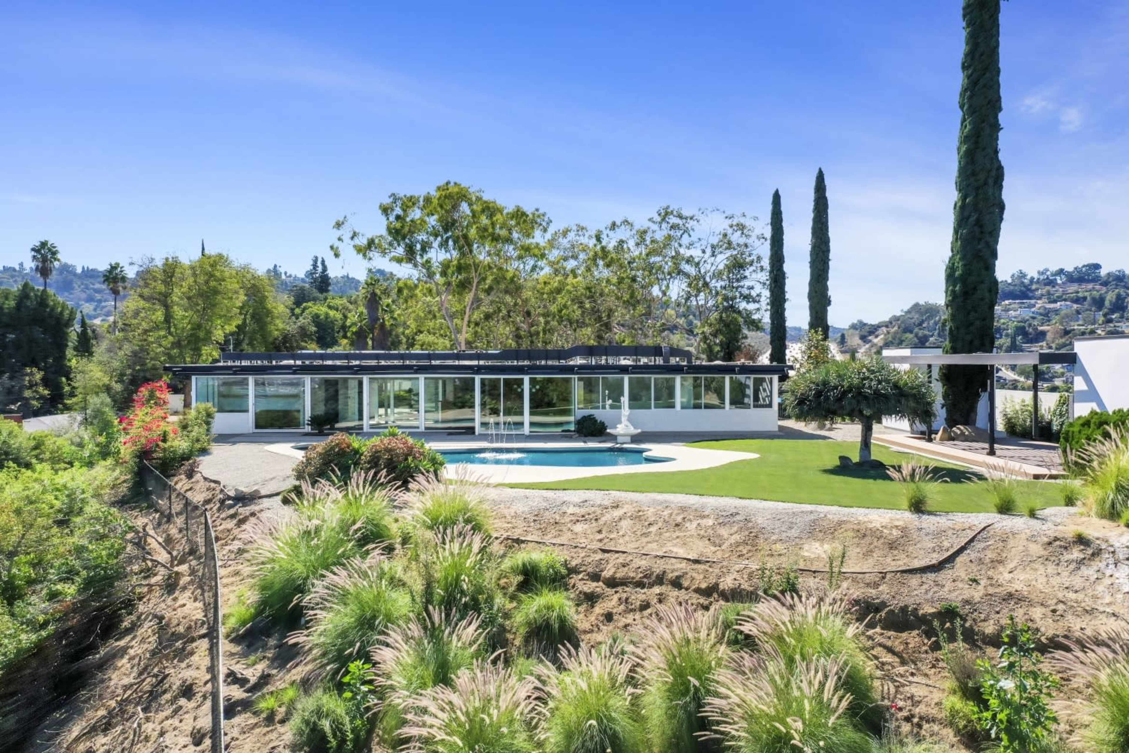 A modern house with large windows surrounds a swimming pool, set against a landscaped garden featuring tall cypress trees and ornamental grasses.
