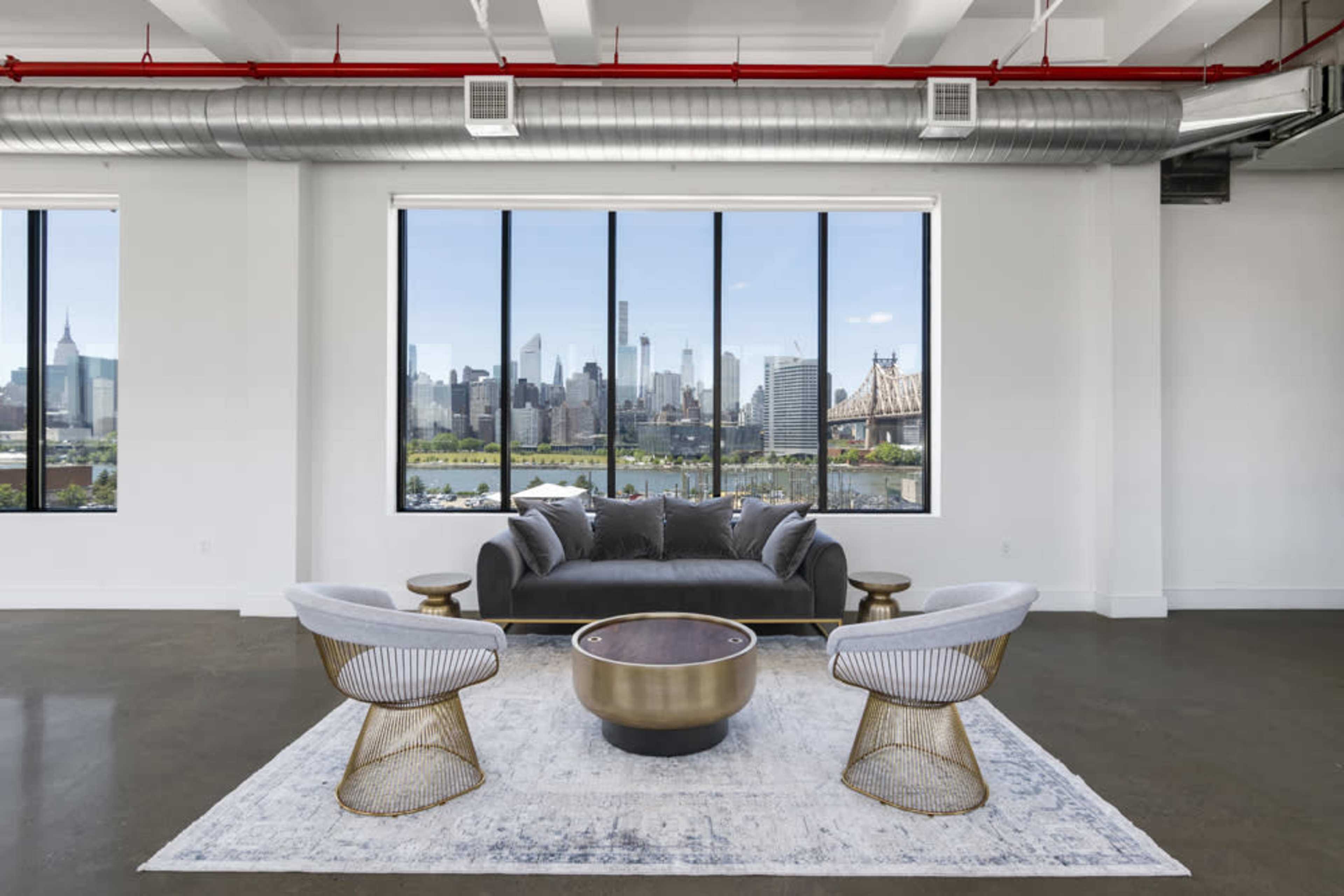 A modern living space with a gray sofa, two armchairs, and a circular coffee table, featuring large windows that overlook a city skyline and body of water.