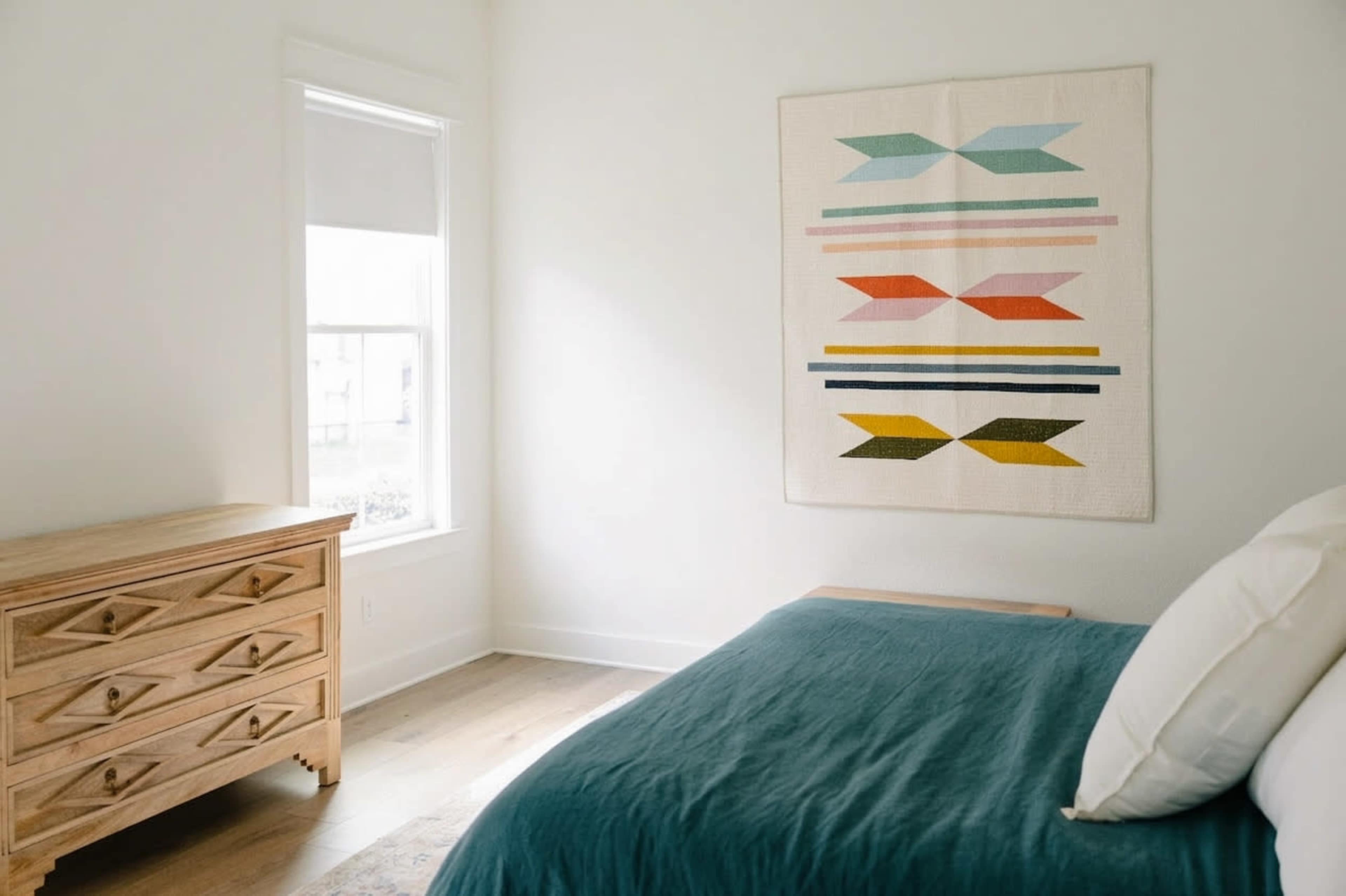 The image shows a bright bedroom featuring a wooden dresser, a bed with a blue cover, and a colorful quilt hanging on the wall.