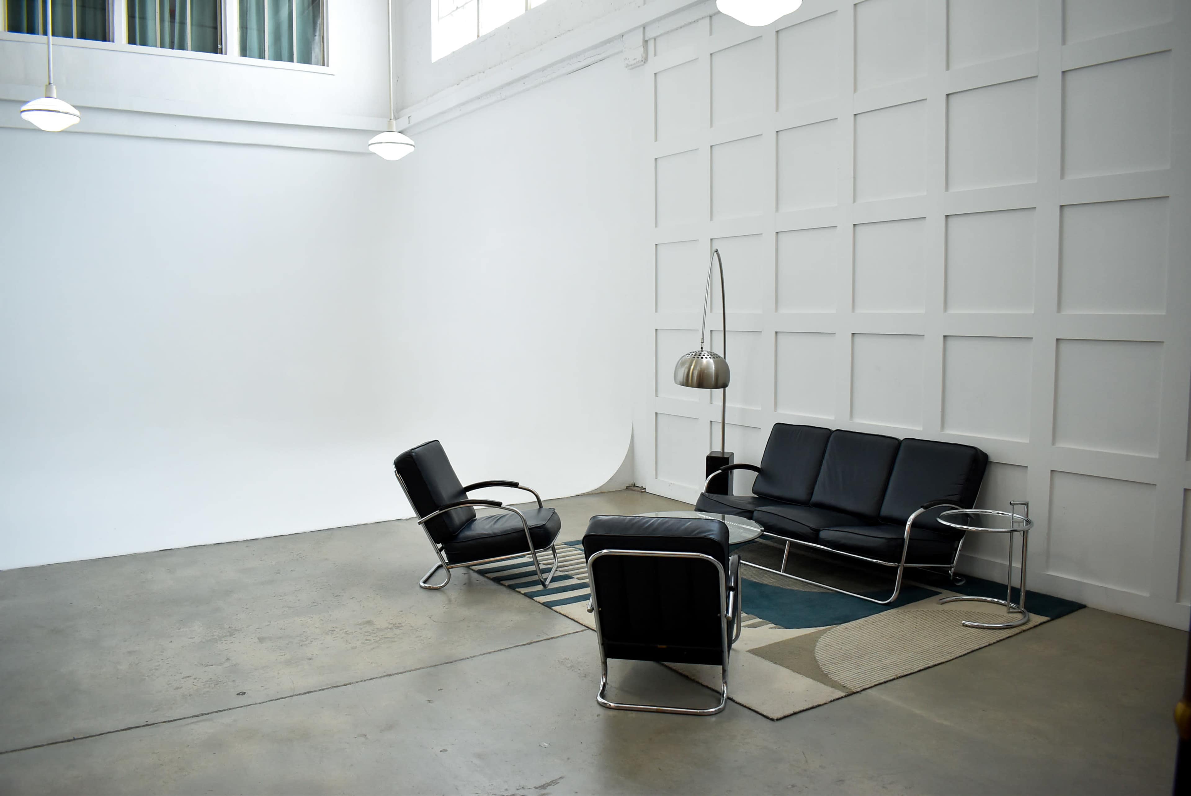 A minimalist seating area with black leather furniture arranged on a patterned rug in a spacious, well-lit room with white walls.