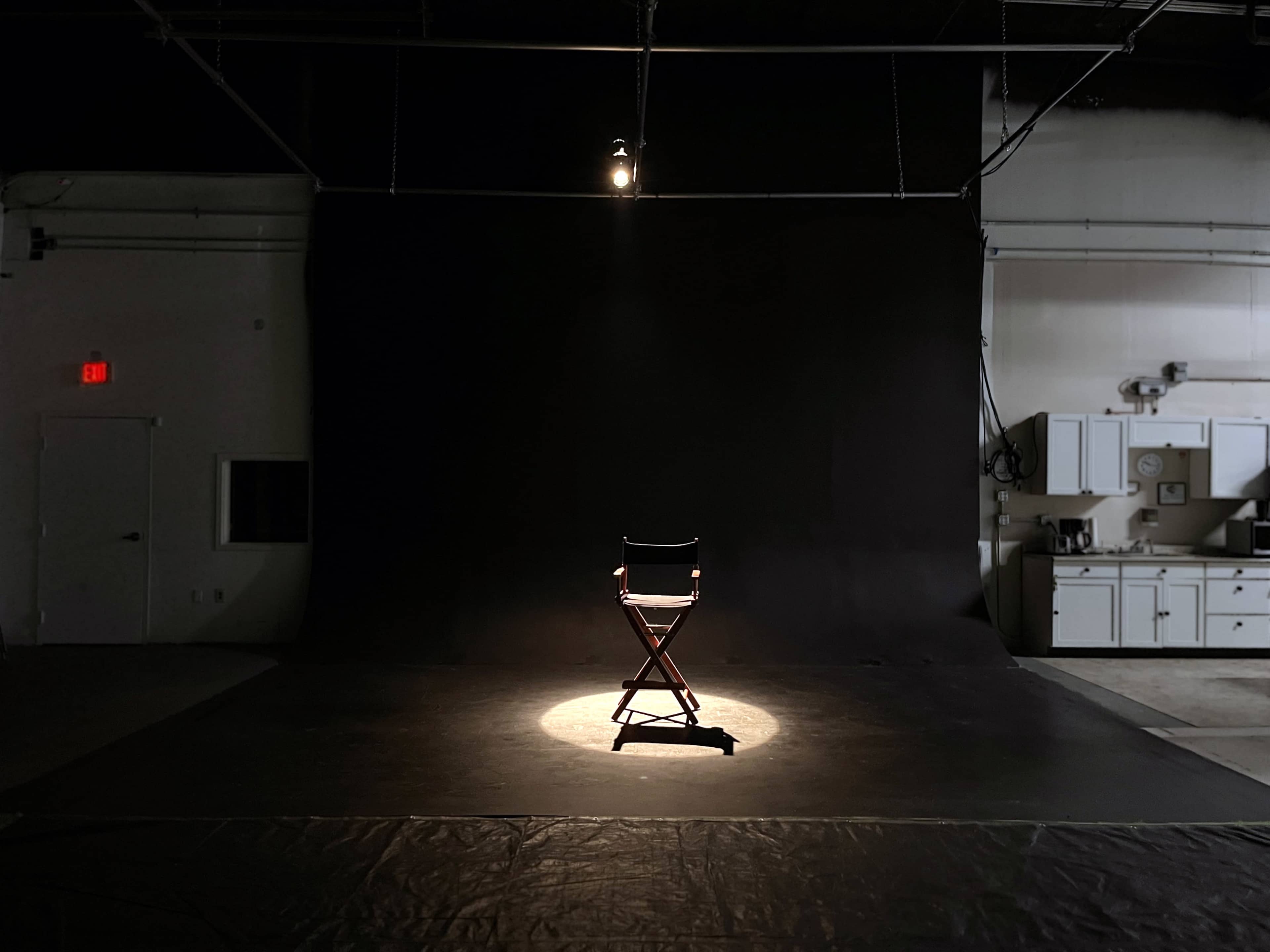 A single director's chair is illuminated by a spotlight on a dark stage with a black backdrop.