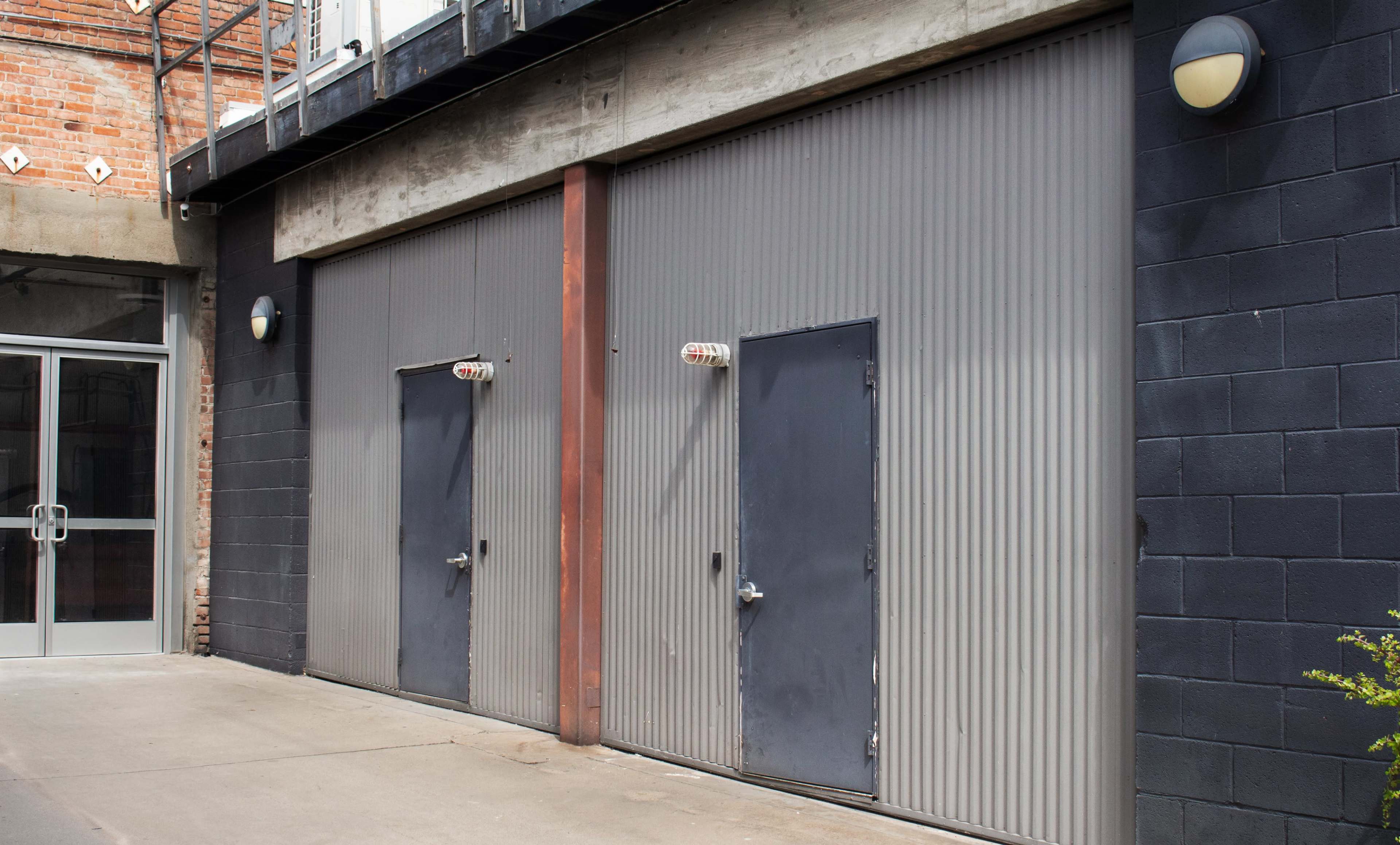 The image shows a brick and concrete building facade featuring two gray metal doors with vertical paneling, positioned side by side against a dark wall.