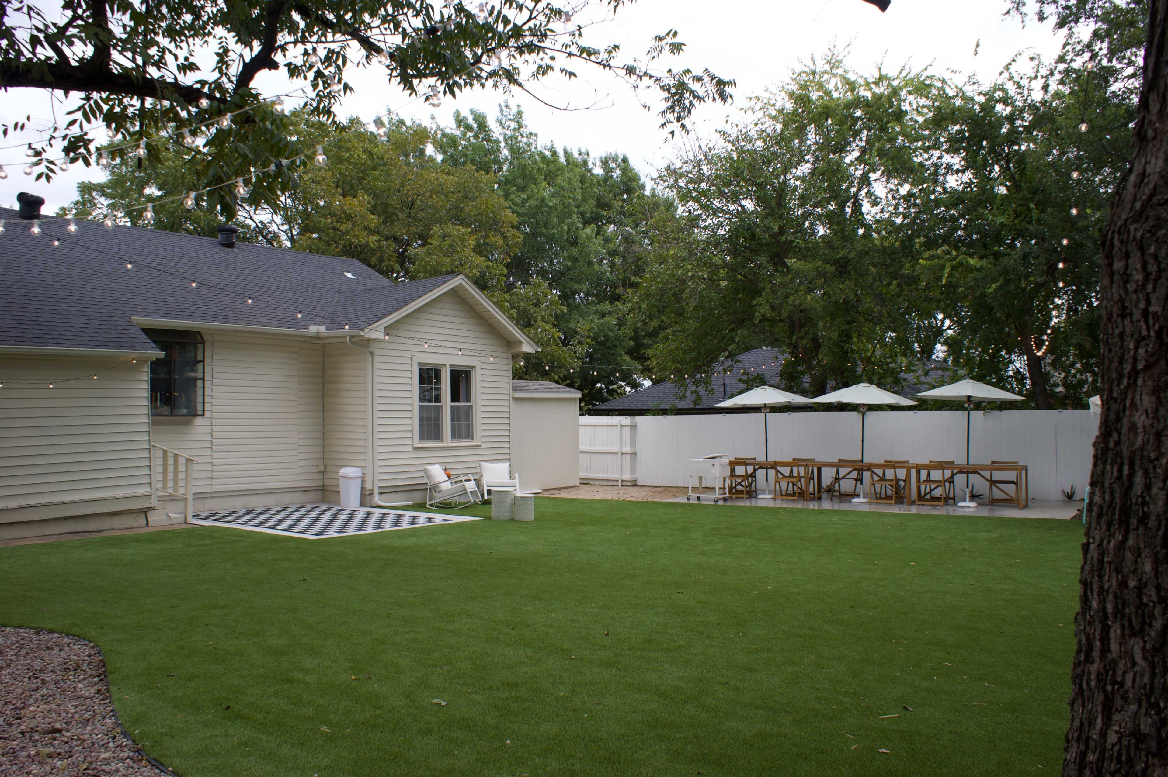 The image shows a backyard featuring artificial grass, a patio with a dining table and umbrellas, and a house with light-colored siding.