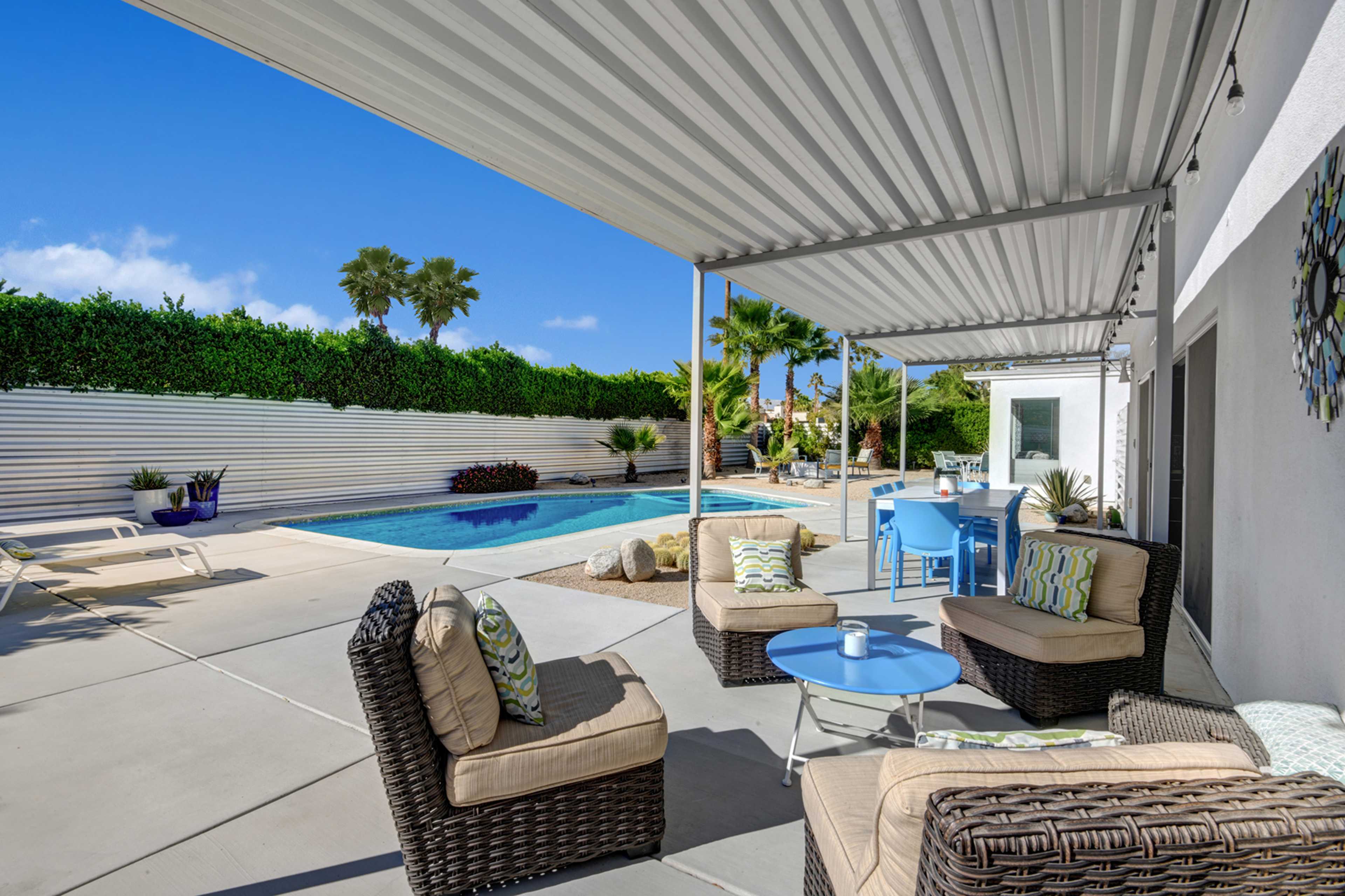 A patio features comfortable seating arrangements and a swimming pool, surrounded by greenery and a bright sky.