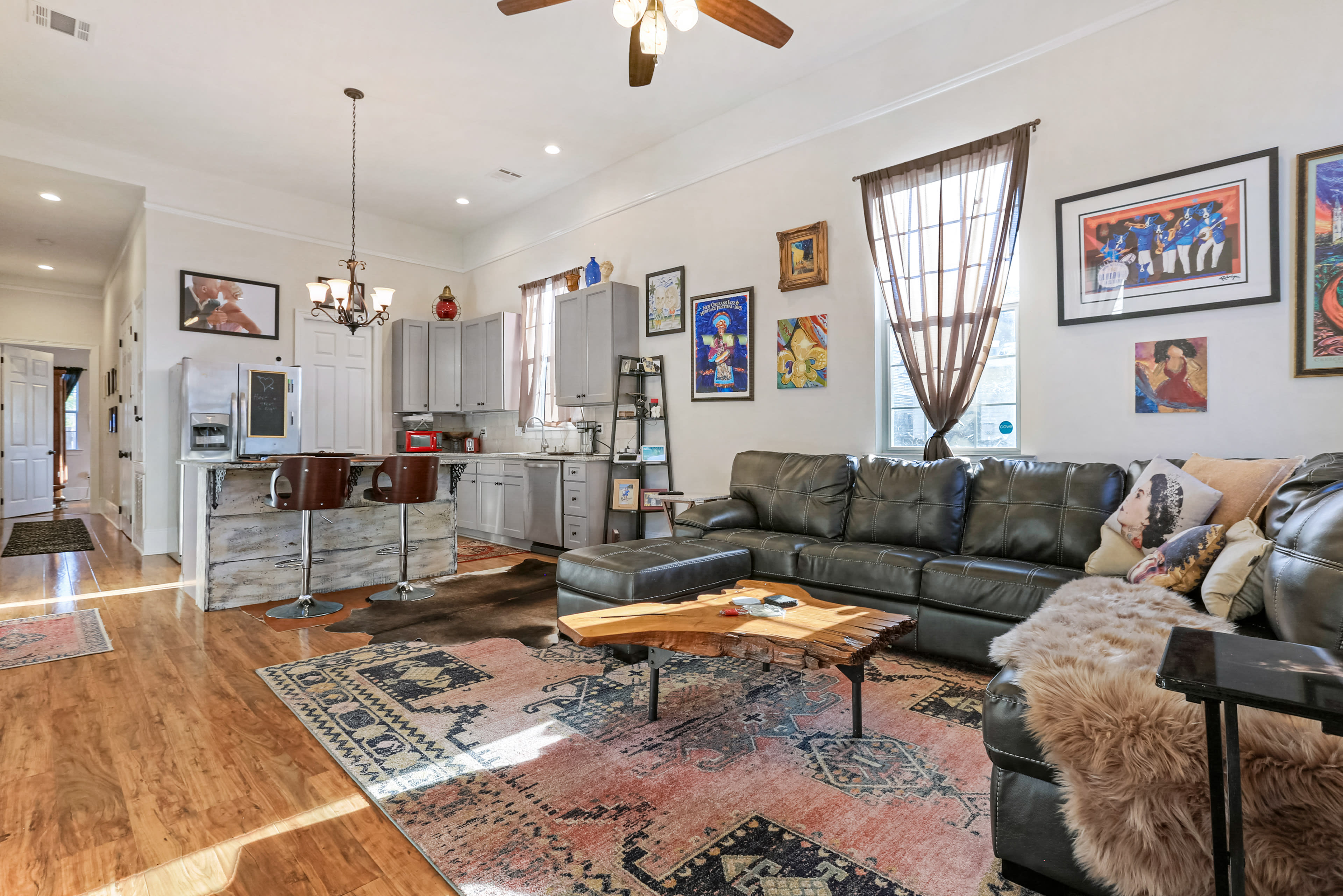 The living room features a leather sectional sofa, a wooden coffee table, and an open kitchen area with bar stools, adorned with various framed artworks on the walls.