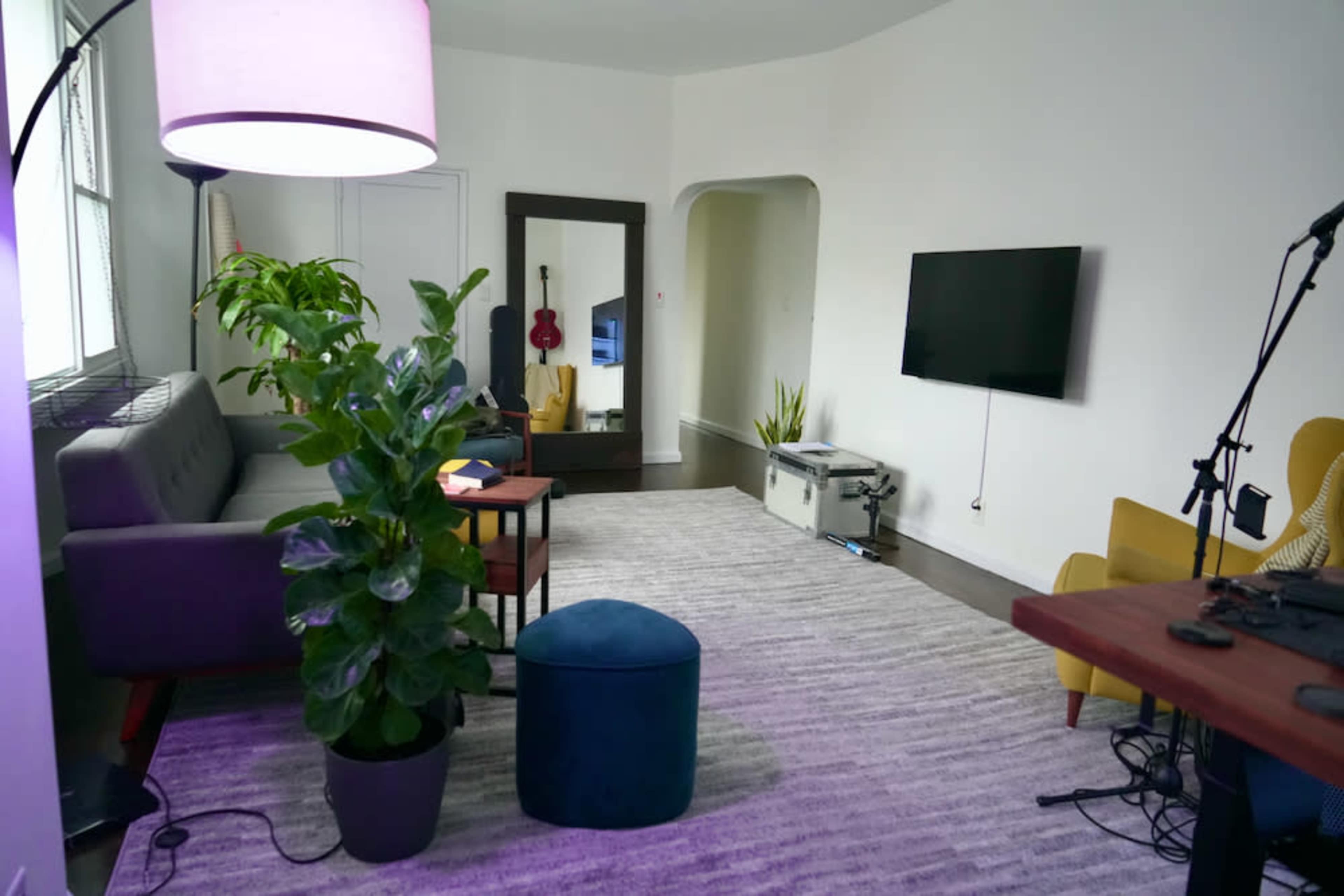 A bright living room with a gray sofa, a green plant, a large mirror, a TV mounted on the wall, and a small blue ottoman on a textured rug.