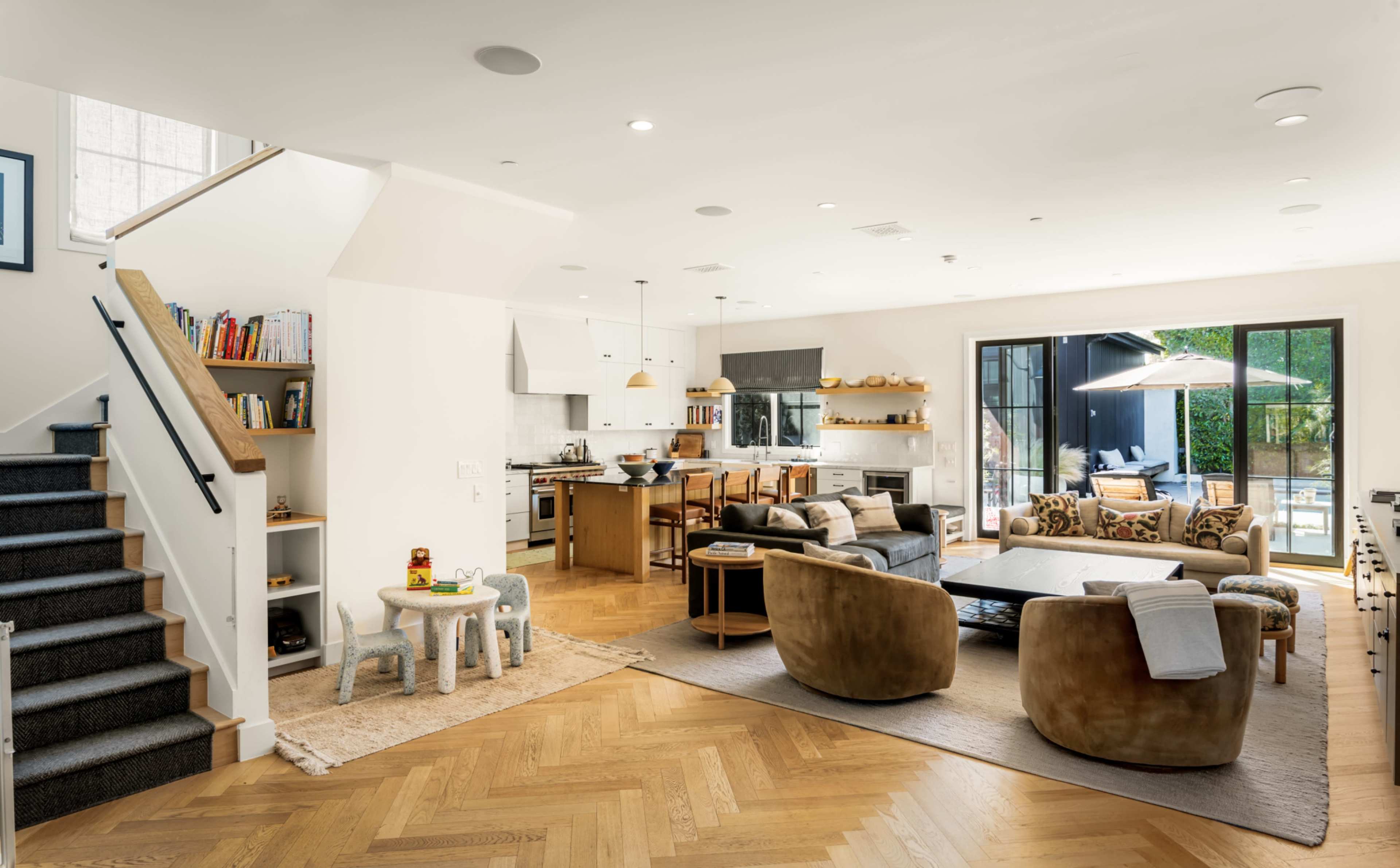 The image shows a modern open-concept living space featuring a kitchen, dining area, and seating area with large windows that open to an outdoor patio.