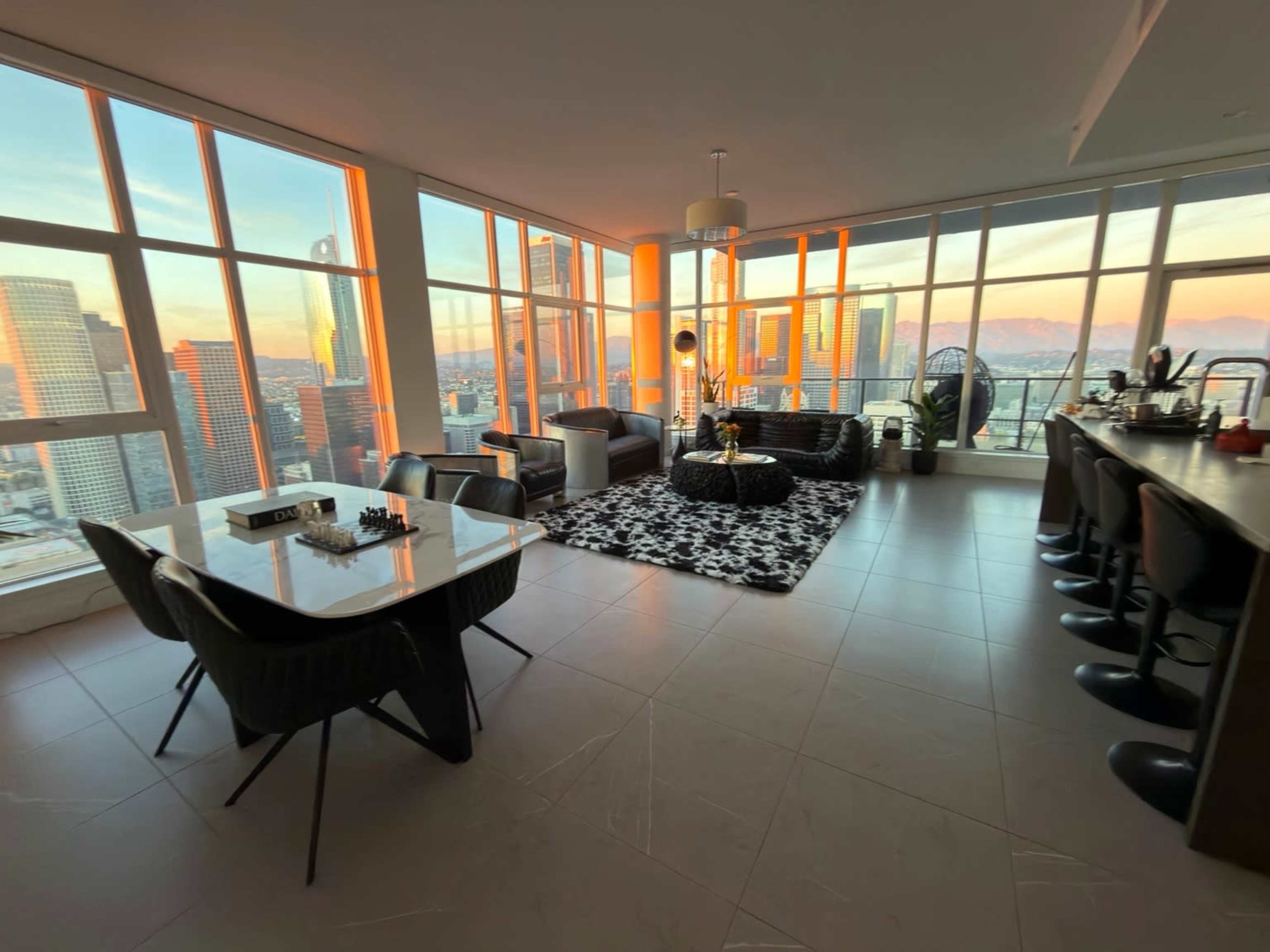 The image shows a modern, stylish apartment with large windows offering a view of a city skyline at sunset, featuring a dining area, living space with a black couch, and a bar with high stools.