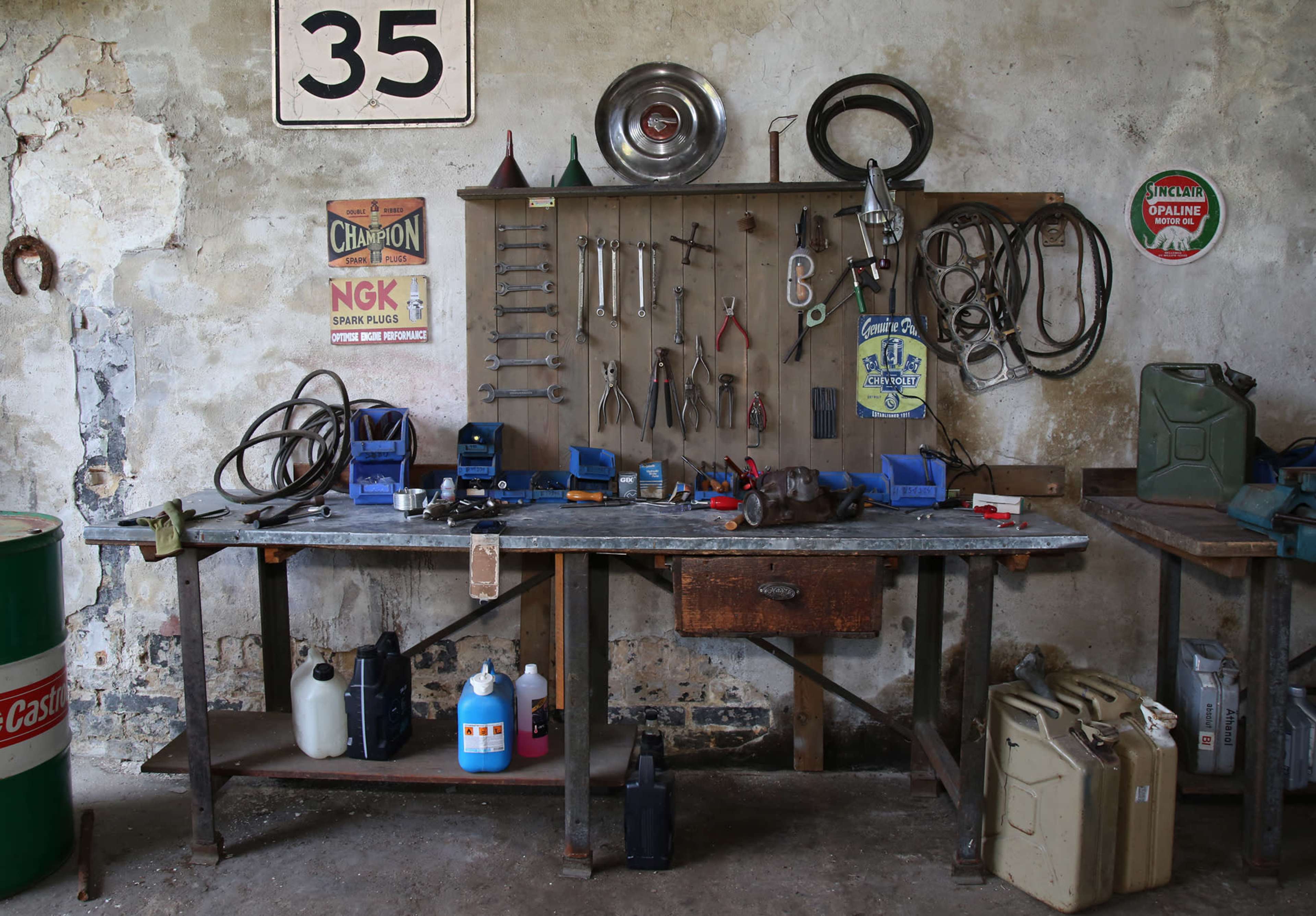 A cluttered workbench features various hand tools, a wall-mounted tool organizer, and containers in a rustic garage setting.