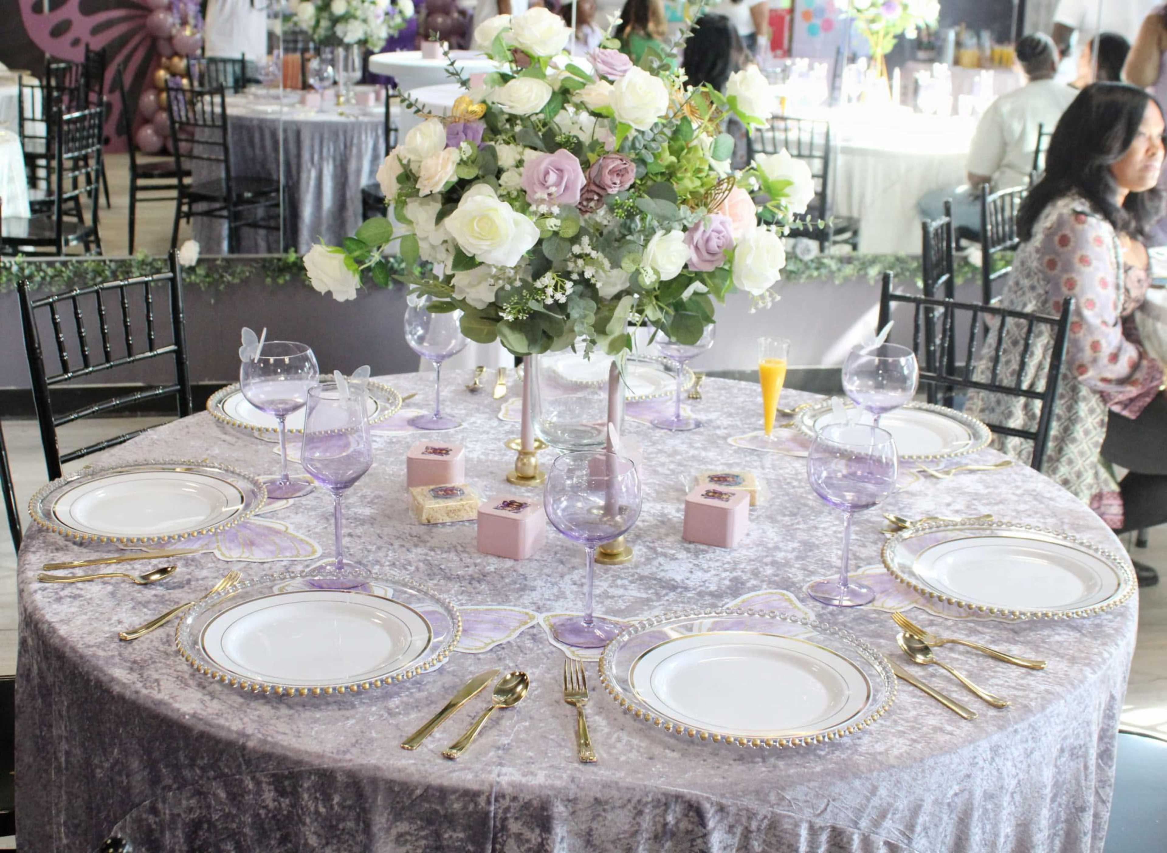 A round dining table is elegantly set with fine china, glassware, and a floral centerpiece, surrounded by guests in a decorated venue.