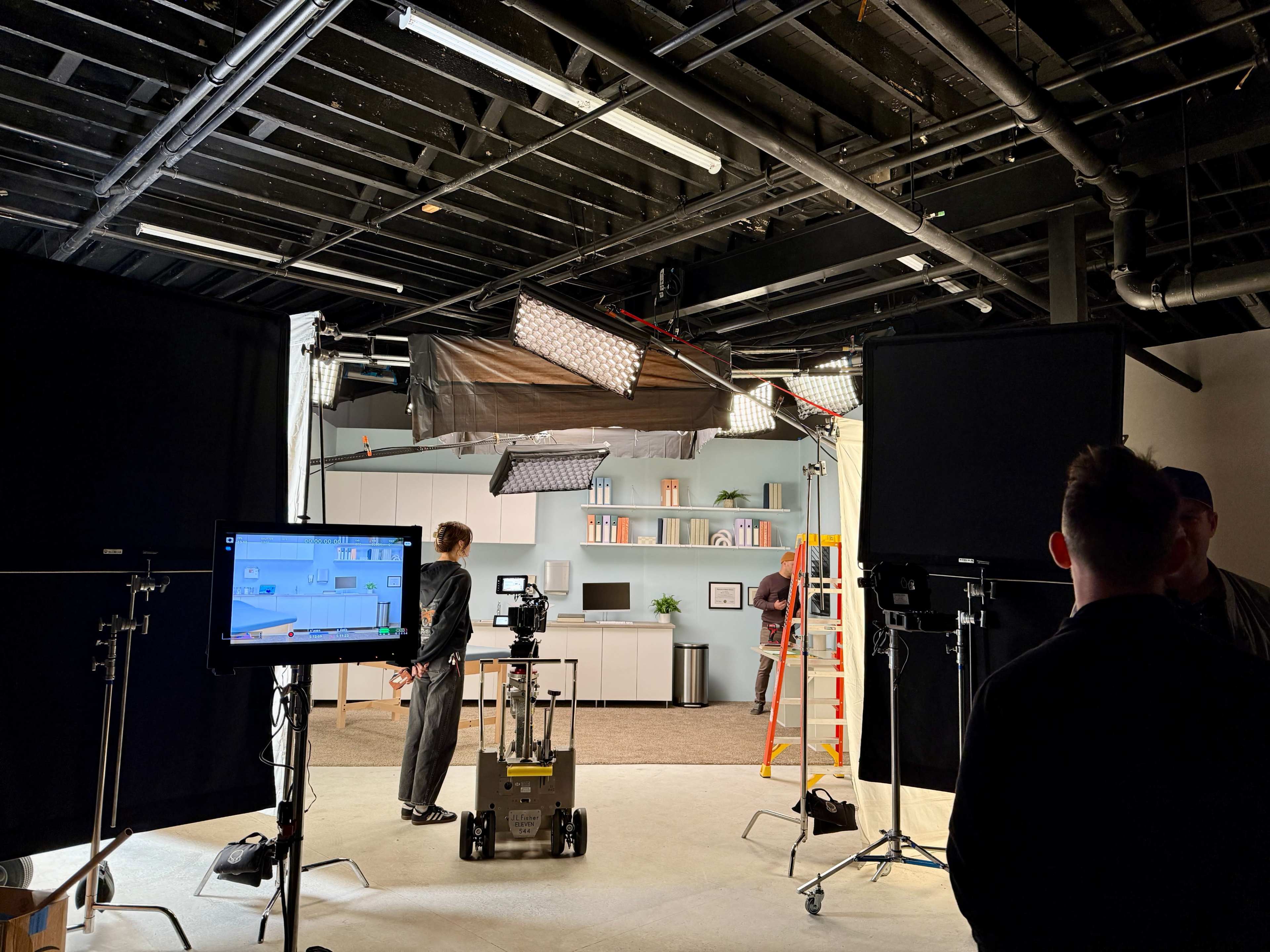 A modern studio setup features equipment and crew preparing for a filming session, with a backdrop of shelves and plants visible.