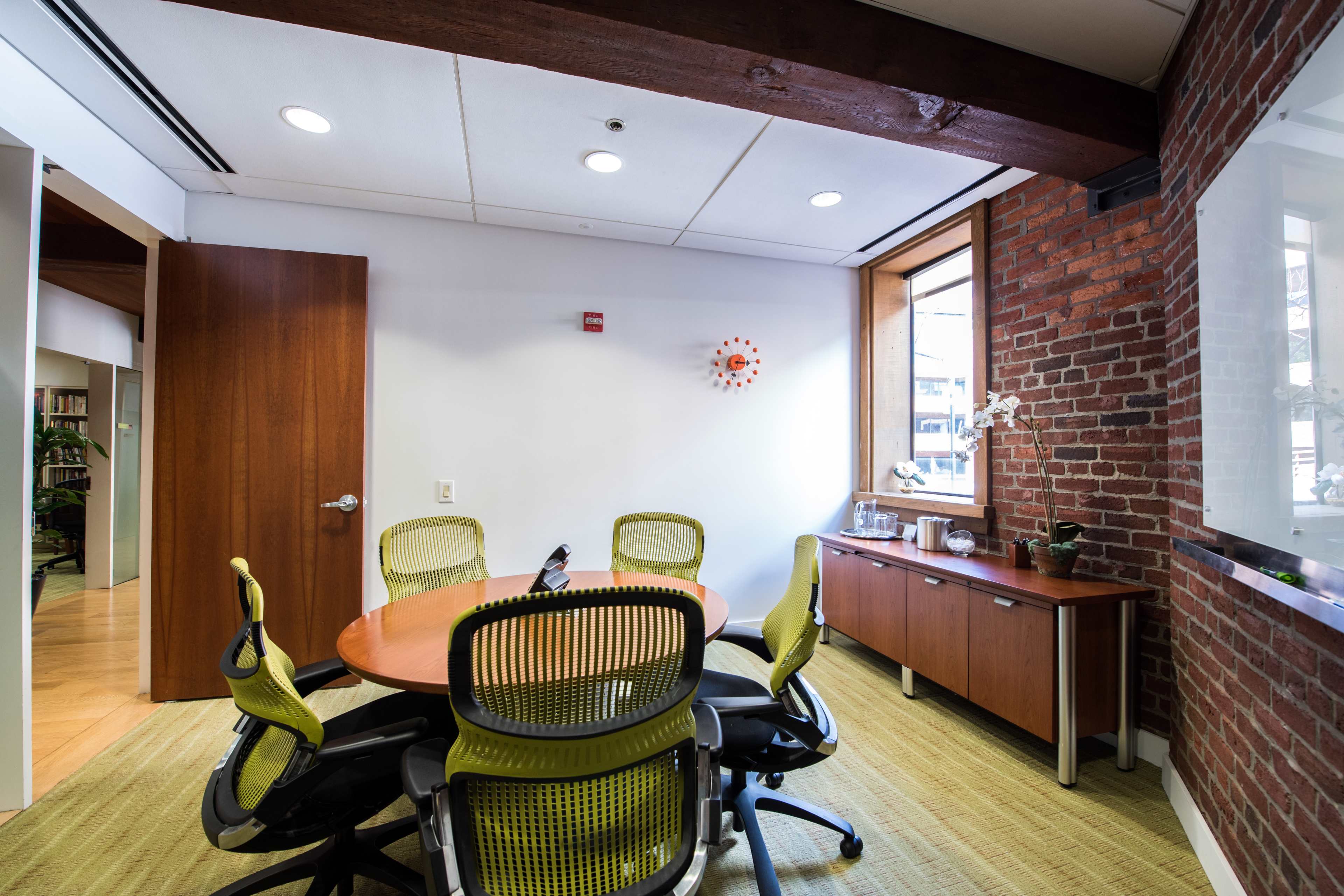 A modern conference room with four green ergonomic chairs around a circular wooden table, featuring a brick wall and a window.
